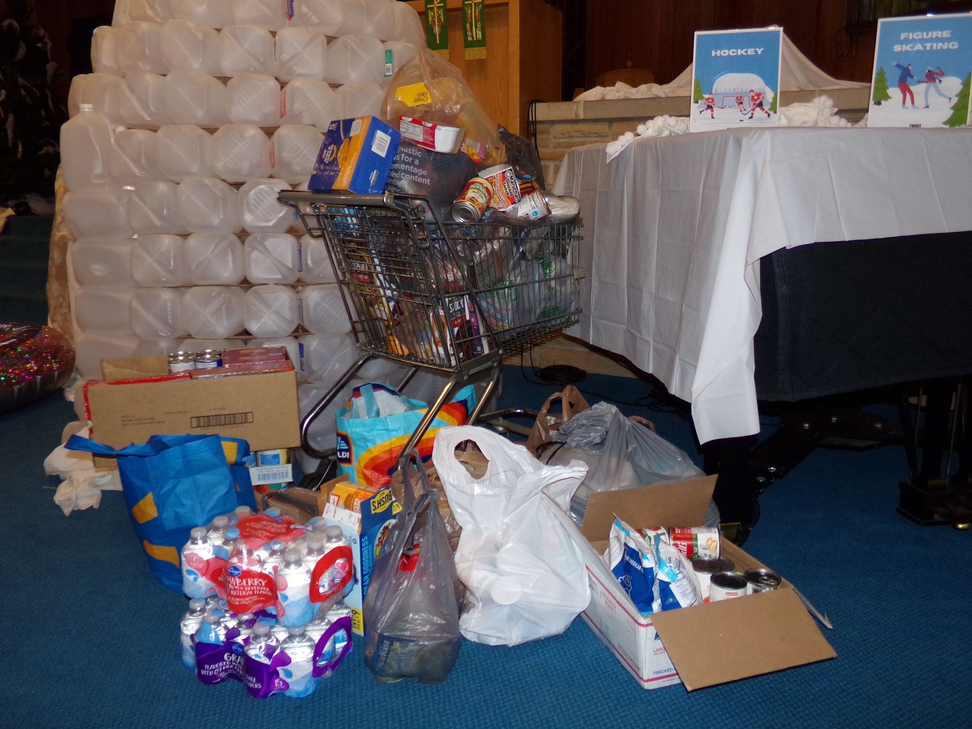 Piles of donated gifts and wrapped packages on a blue-carpeted floor beside a table and balloon display.