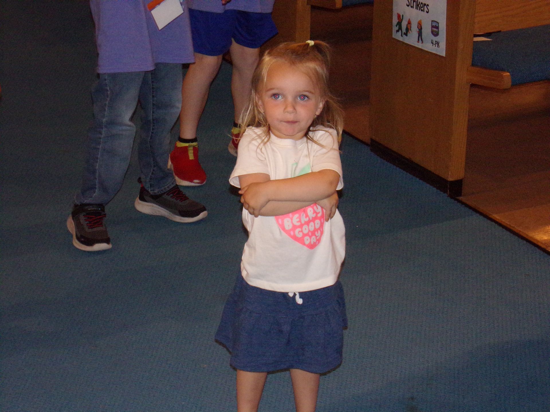 Young child standing with arms crossed in a blue-carpeted room, wearing a white shirt and navy skirt.