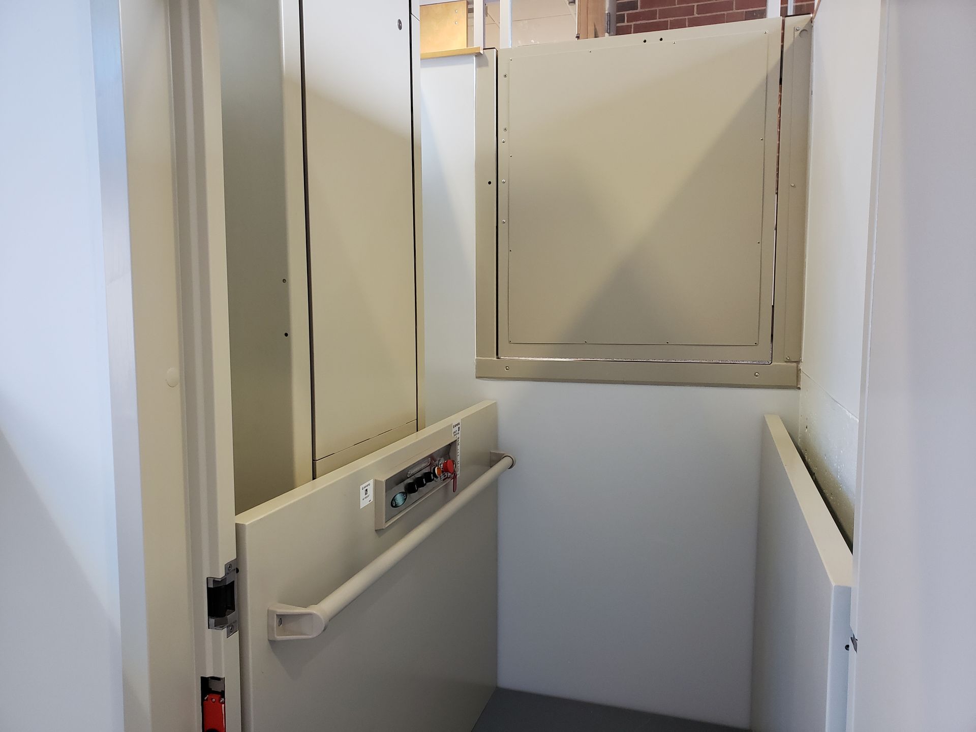 Empty narrow elevator interior with beige walls, handrails, and a closed gate at the back