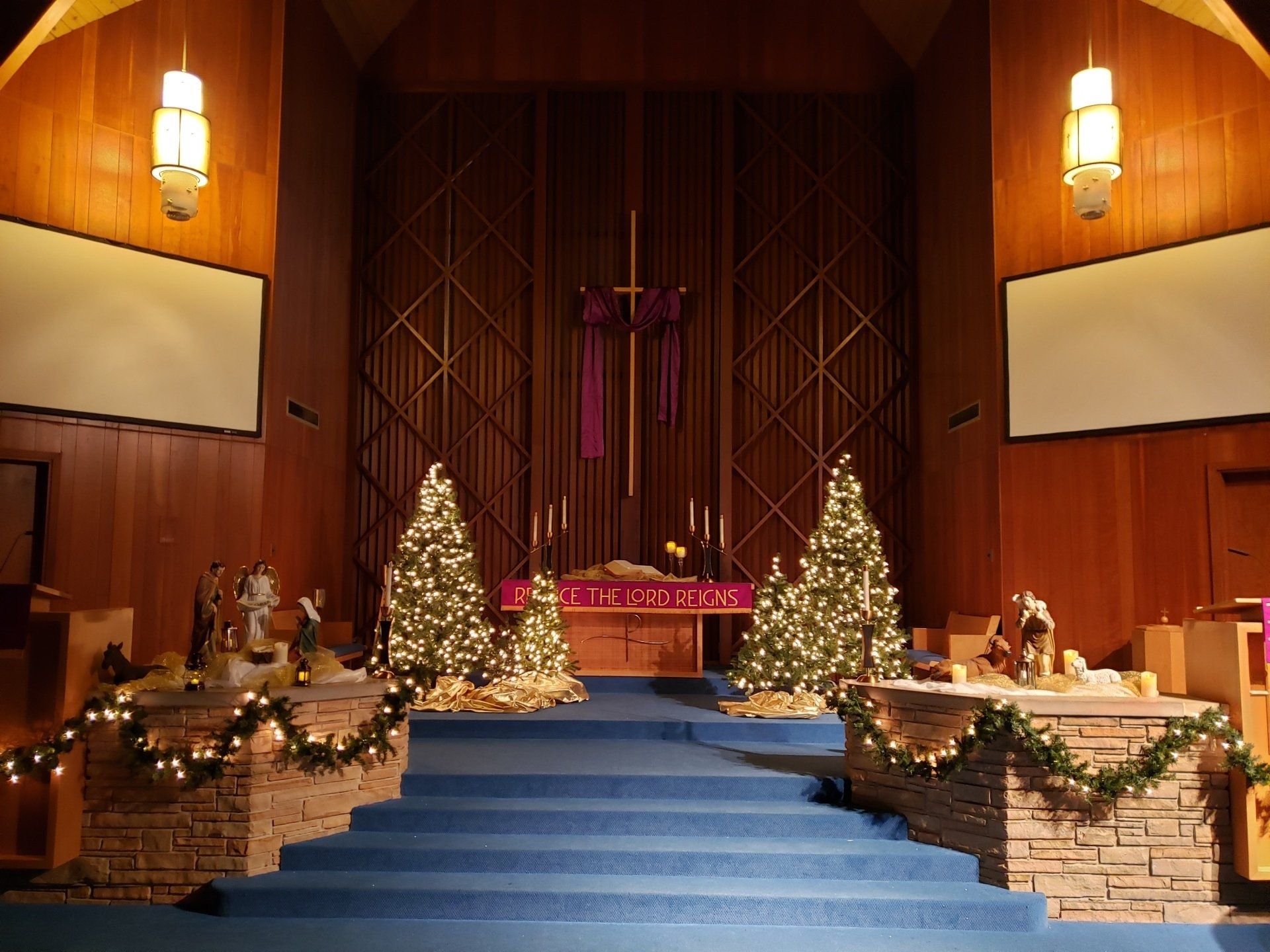 Decorated church altar with blue steps, Christmas trees, and purple Advent wreath in a wood-paneled sanctuary