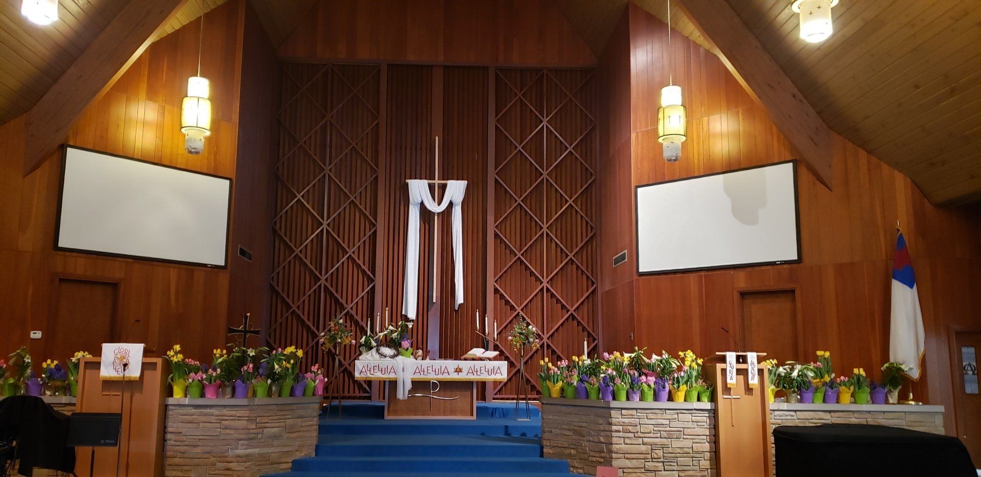 Church altar with blue carpet, floral arrangements, and a central cross under warm wooden walls