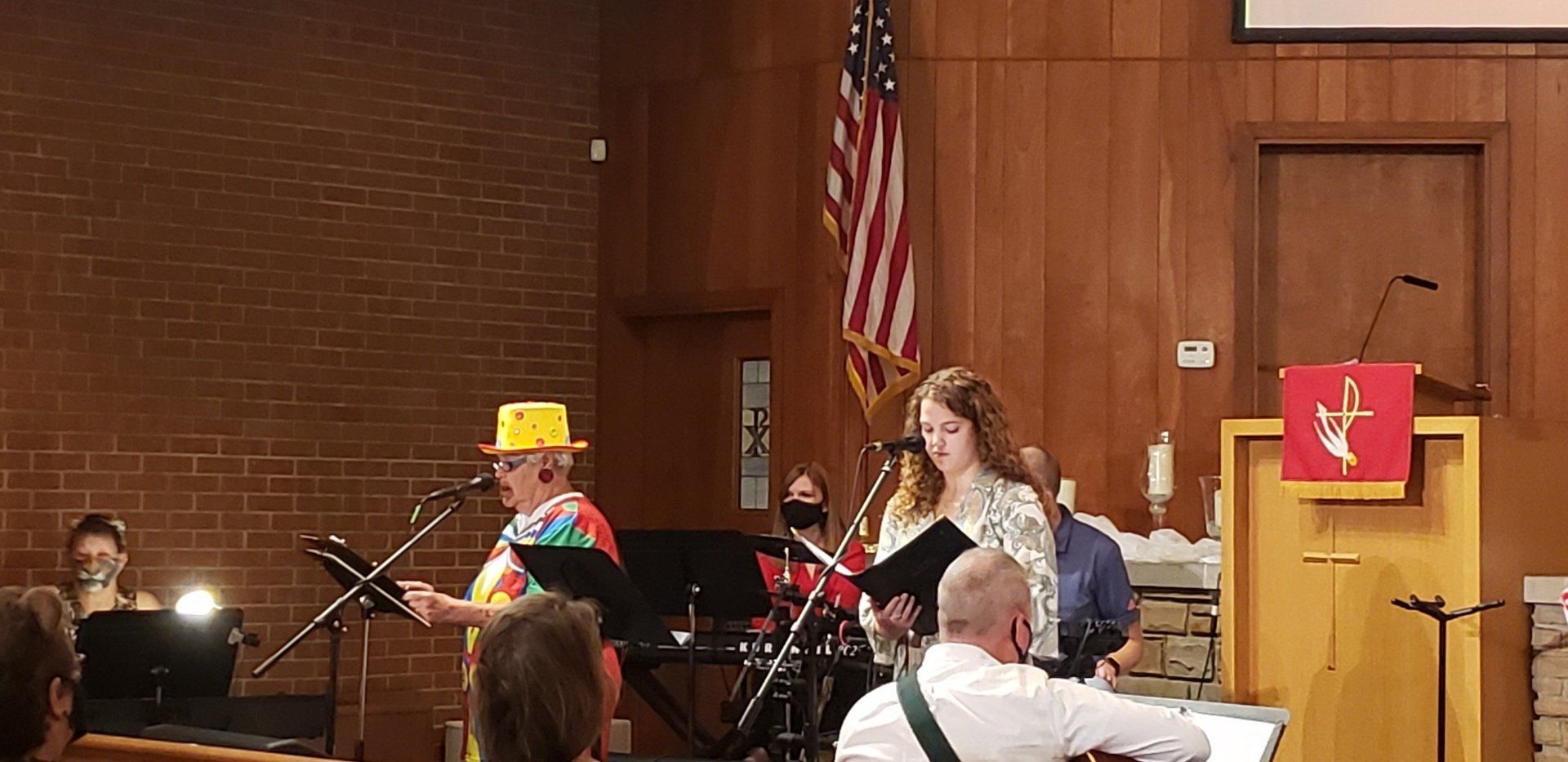Musicians performing on a church stage with music stands, an American flag, and a podium with a red banner