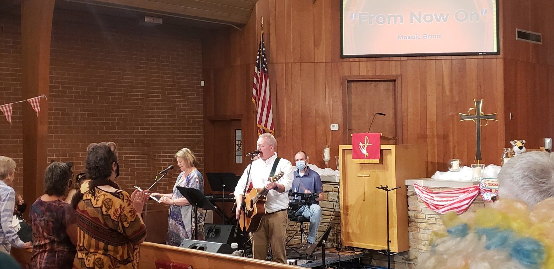 Musicians performing in a church sanctuary with wooden walls, an American flag, and a pulpit beside a decorated altar