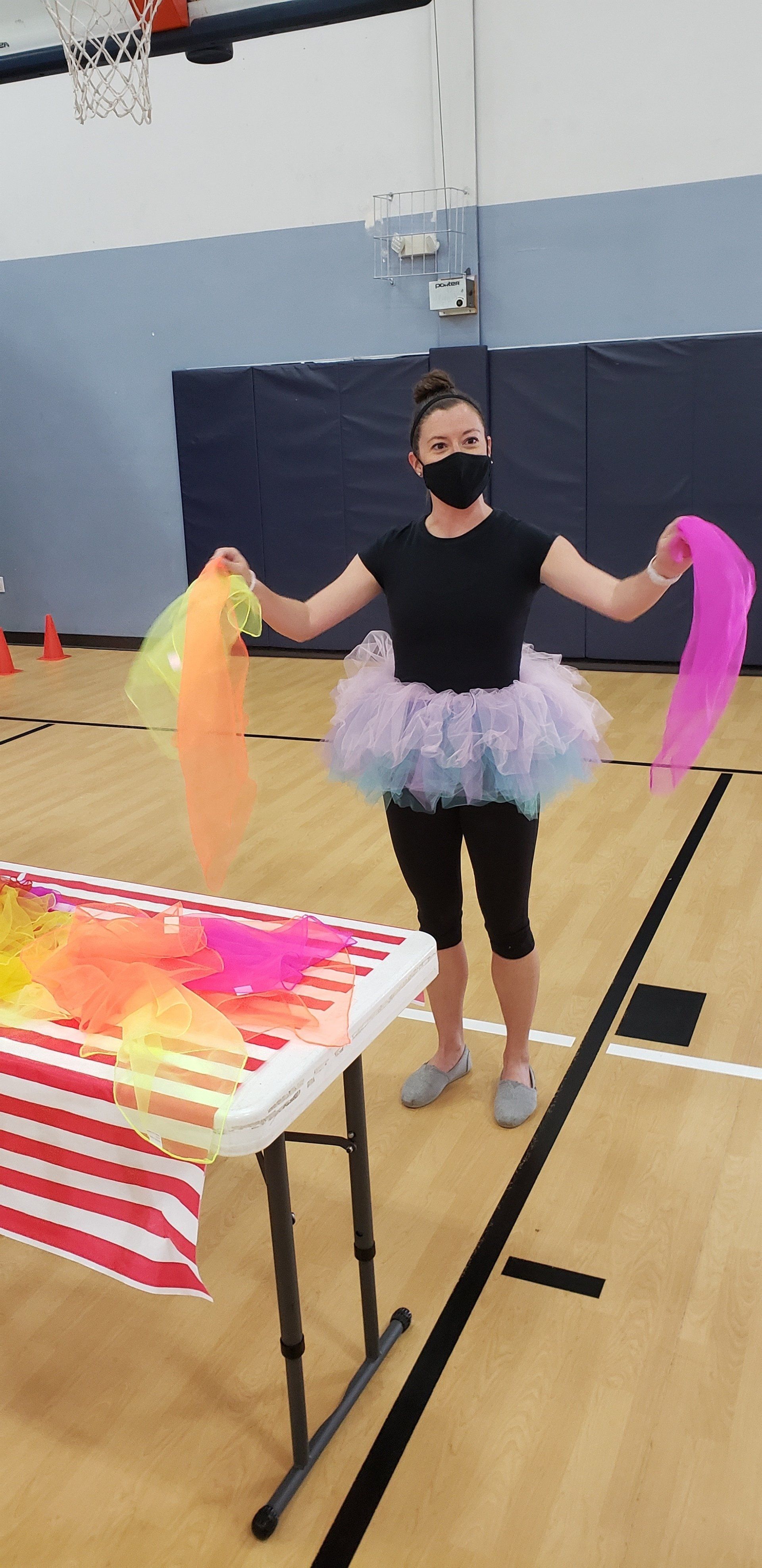 Masked dancer in a gym, wearing a tutu and colorful ribbon streamers with arms outstretched.