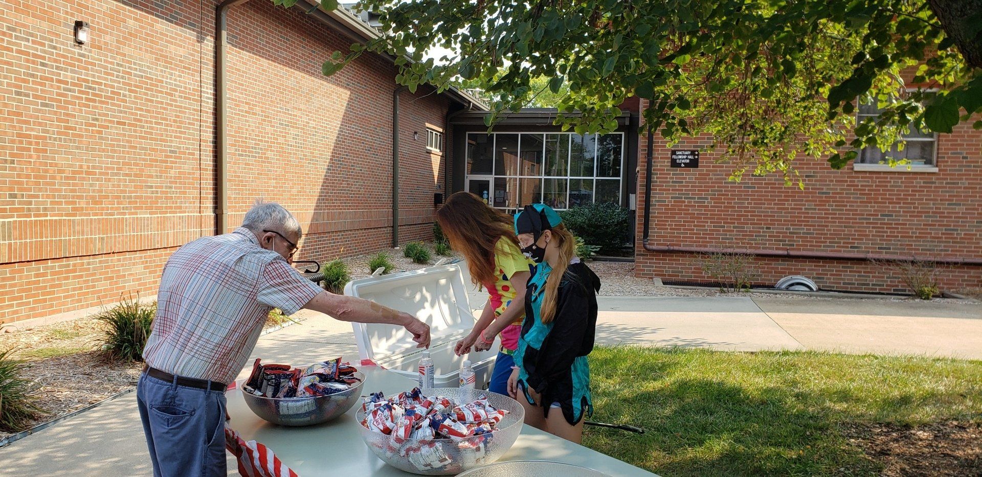 People placing items on a blanket outdoors beside a brick building and trees