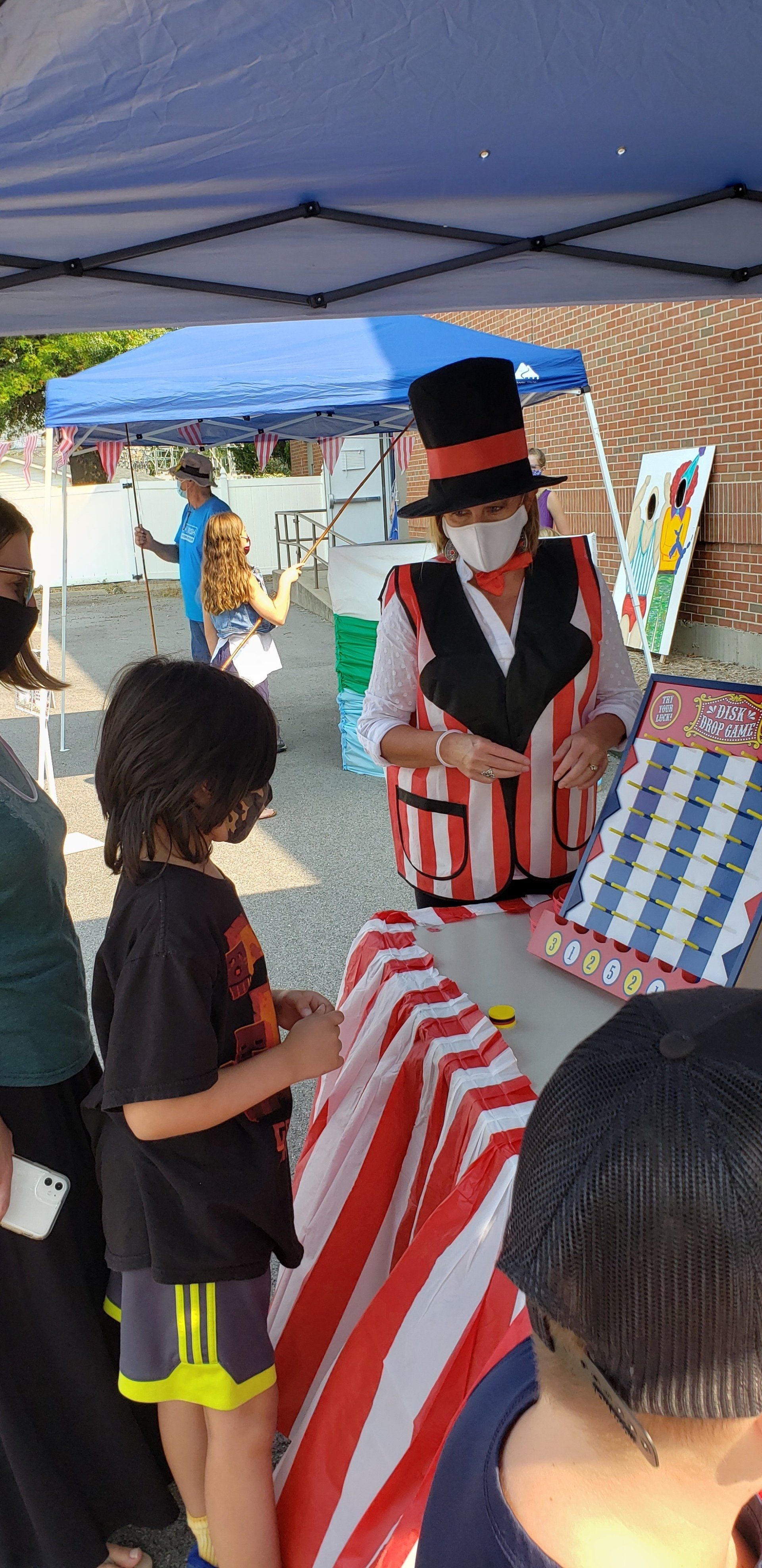 Person in a black top hat stands at a striped booth while a child looks on under a blue canopy.