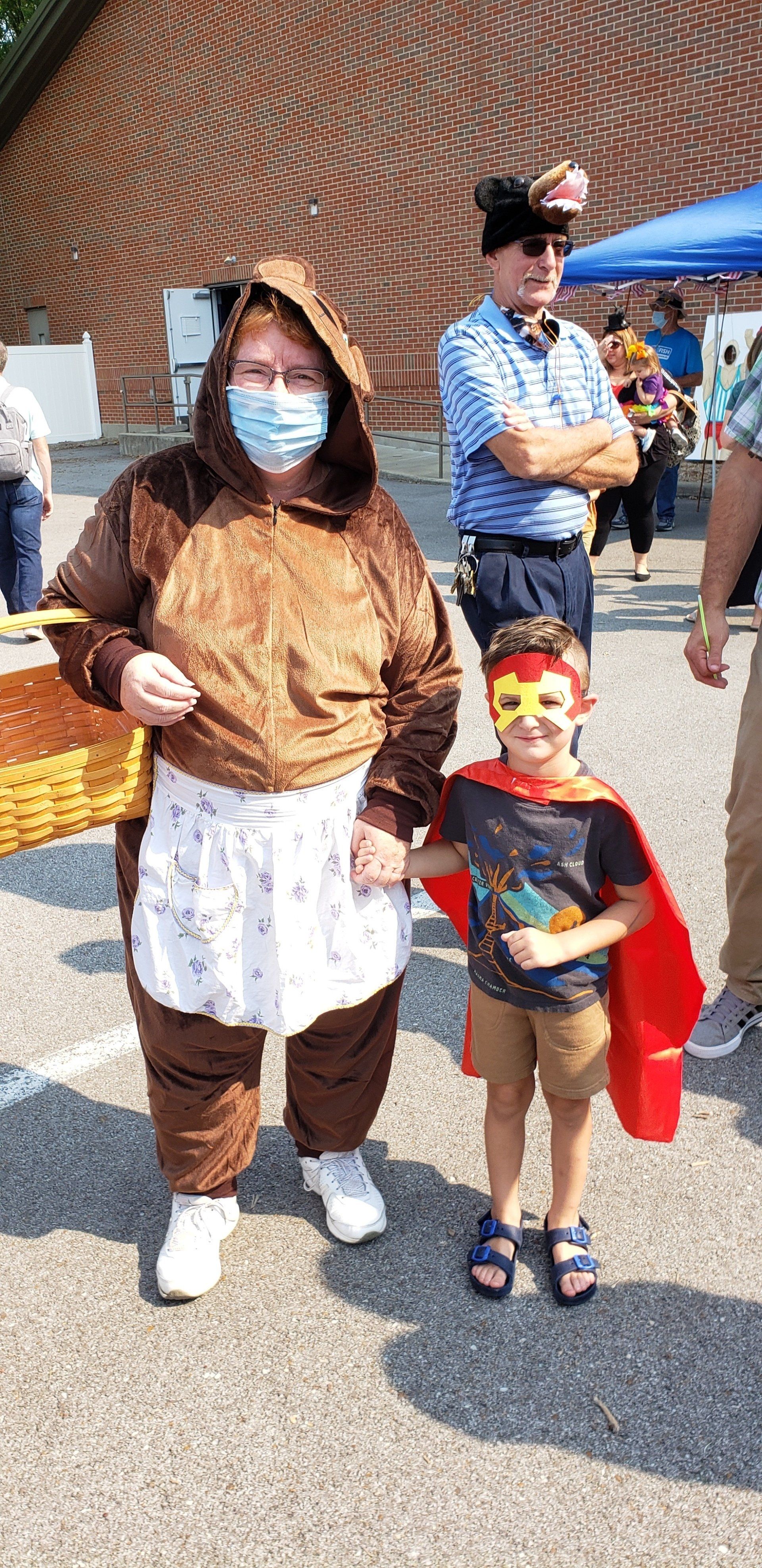 Two costumed children standing outdoors at a festive event, one holding a red cape and mask.