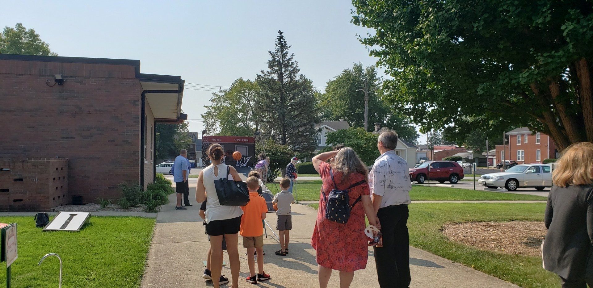People standing on a sidewalk near a brick building, with children and a red car in the background