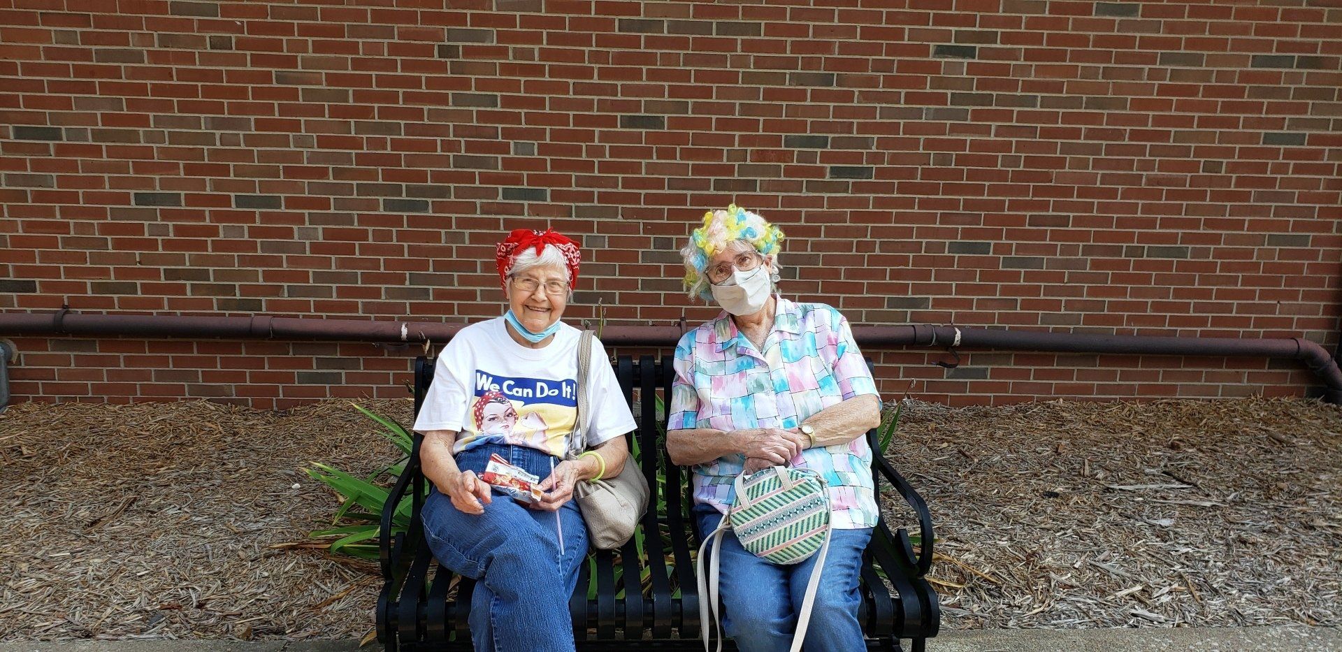 Two seated people in front of a brick wall, wearing colorful patterned clothing and headwear.