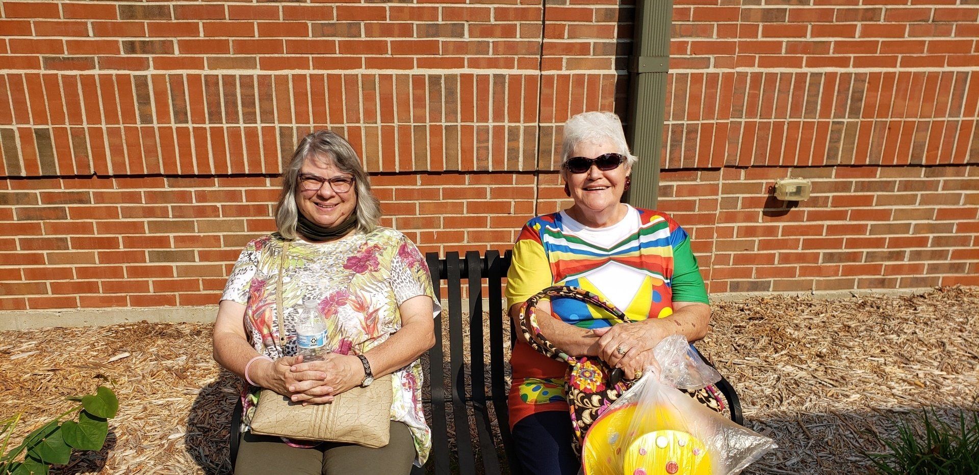 Two people sitting on the grass in front of a brick wall, smiling in bright daylight.