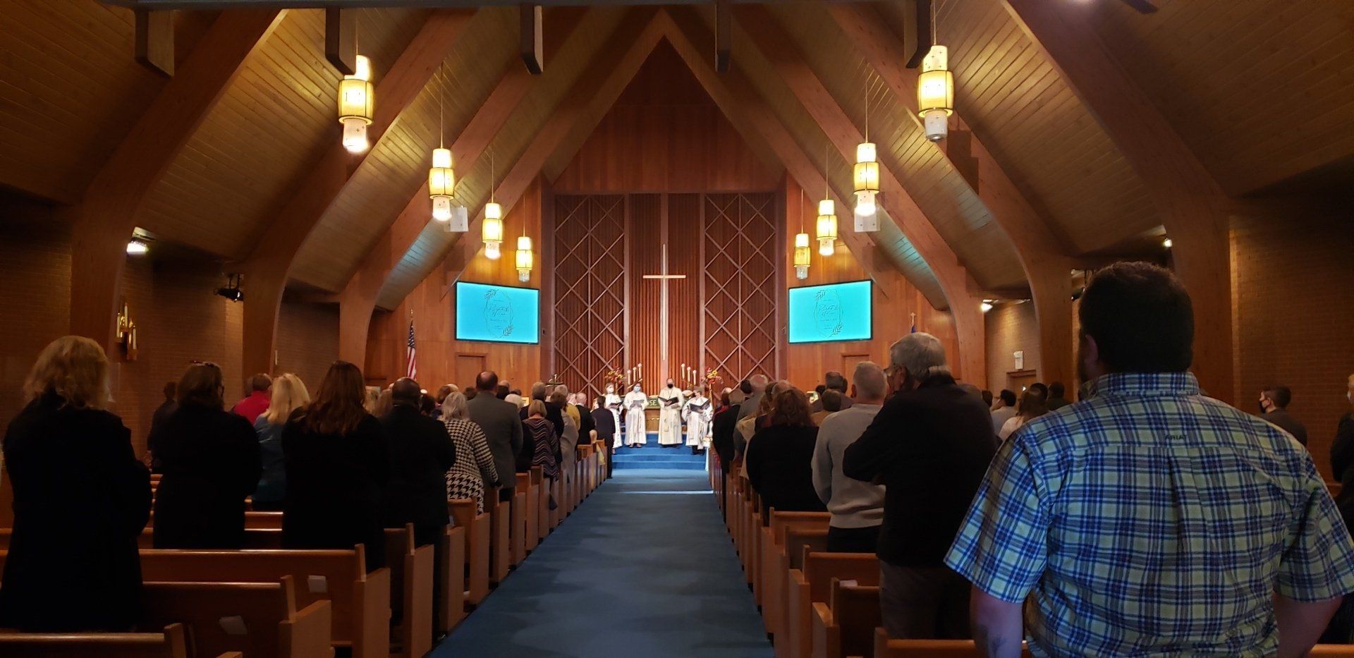 Congregation seated in a wooden church sanctuary, facing a cross and speakers at the front.