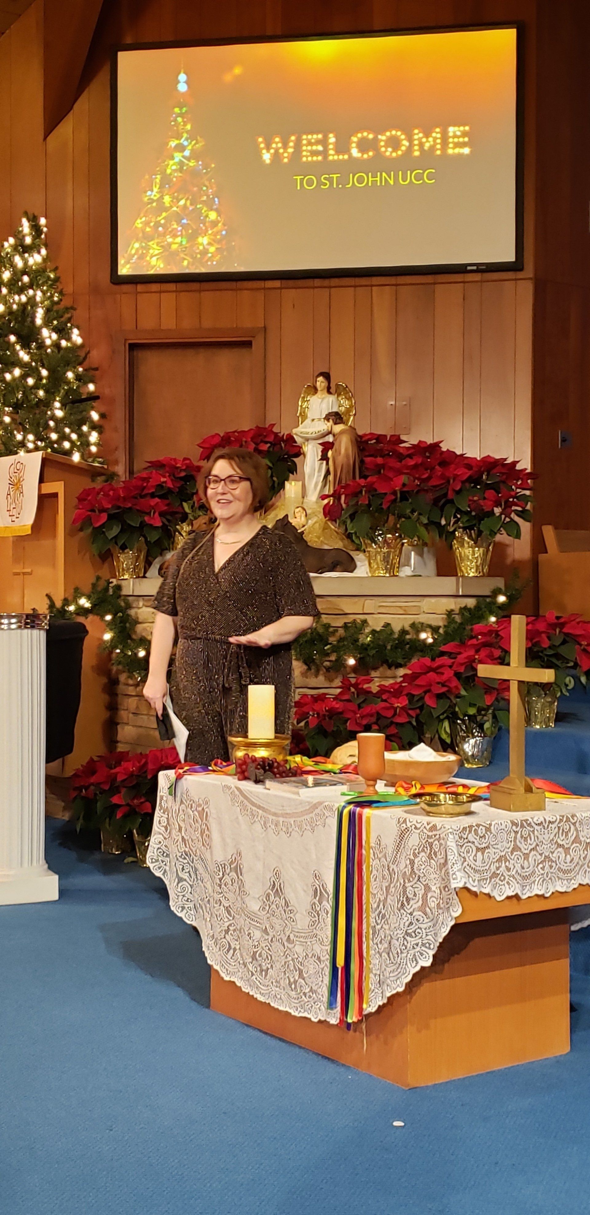 Woman in a black dress stands by a decorated altar with poinsettias beneath a “WELCOME” sign.
