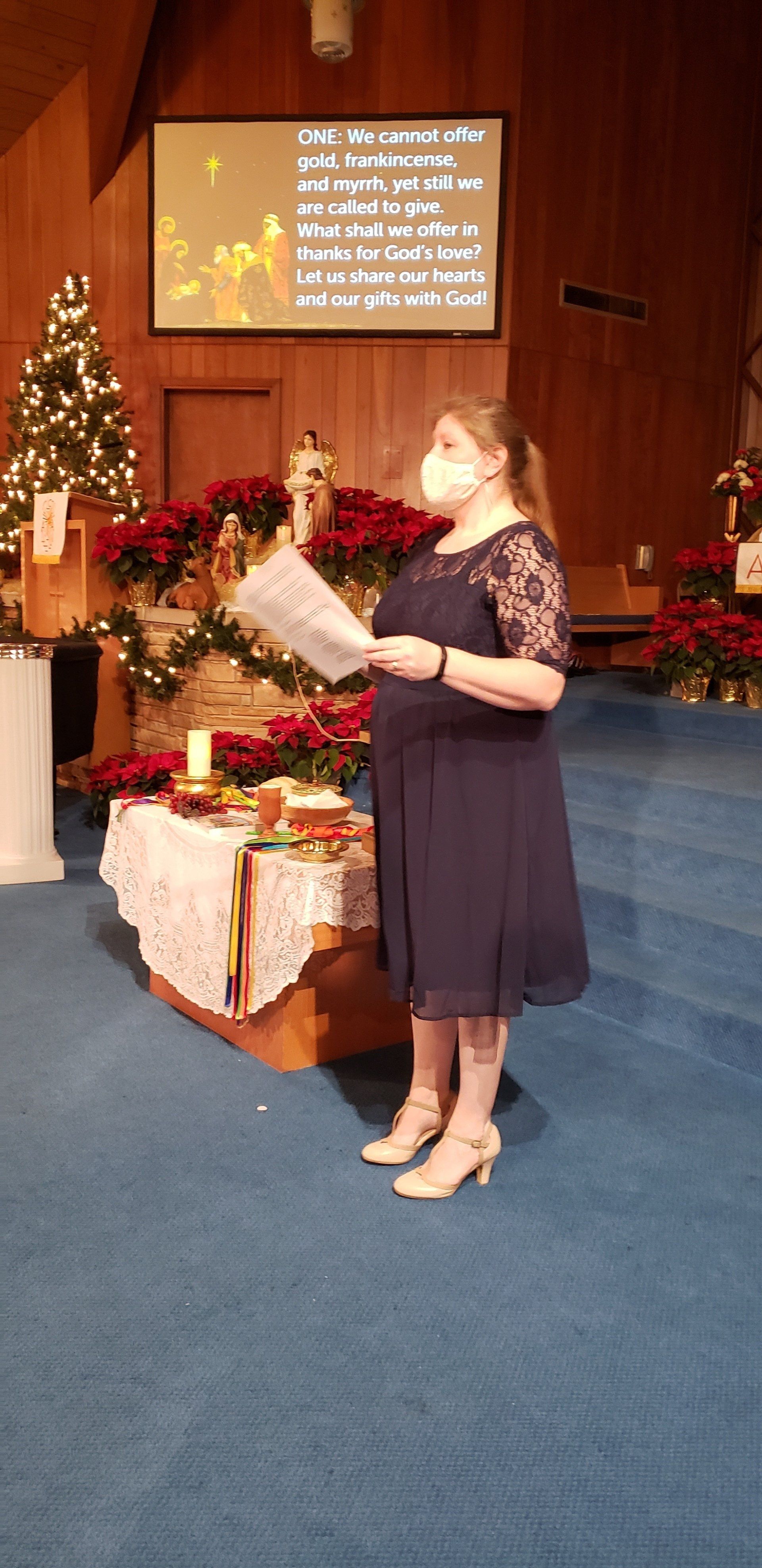 Woman reading from a booklet beside Christmas gifts and a decorated tree on a church stage