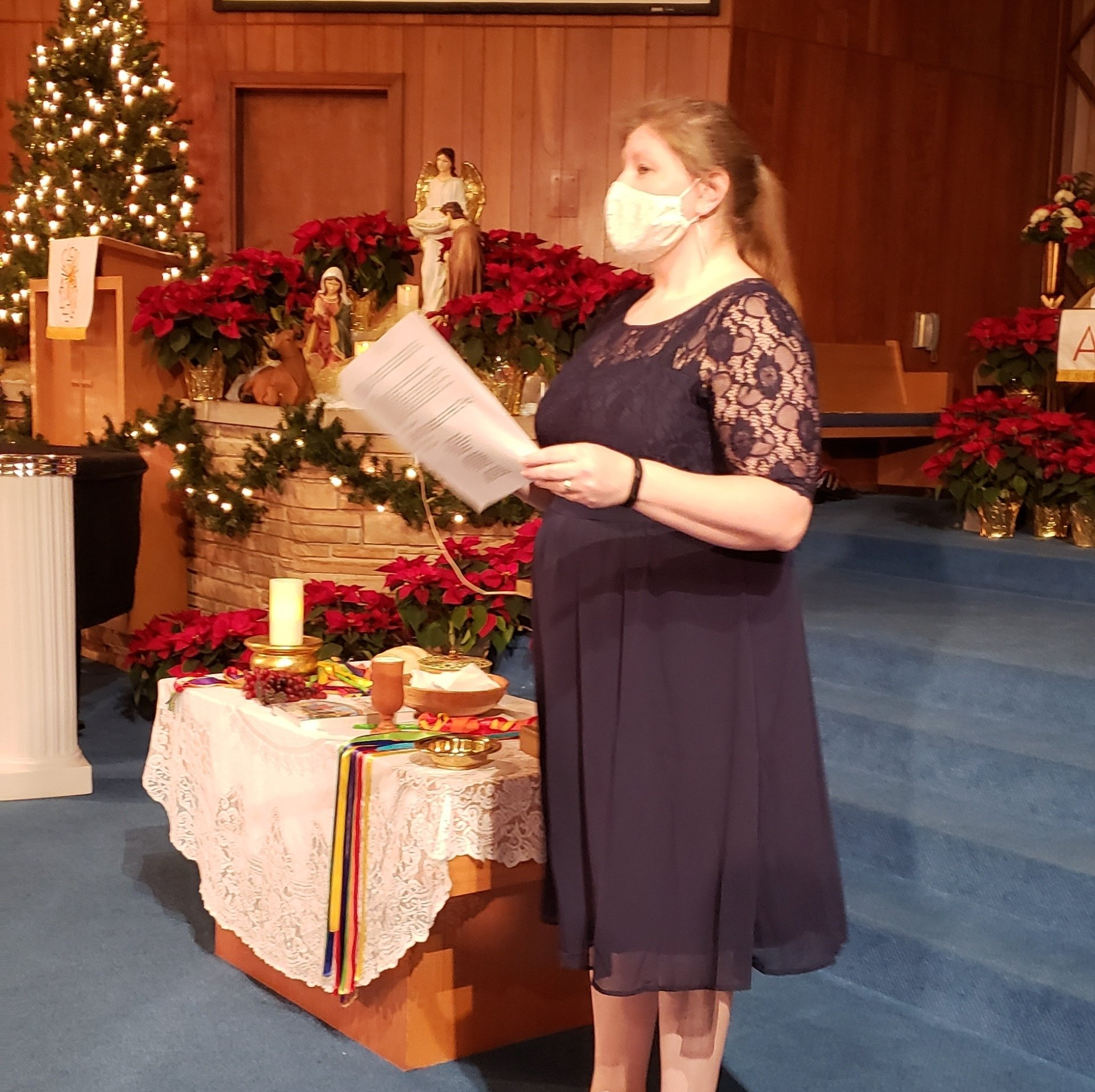 Woman in a mask reading from a paper at a church altar decorated with Christmas lights and poinsettias