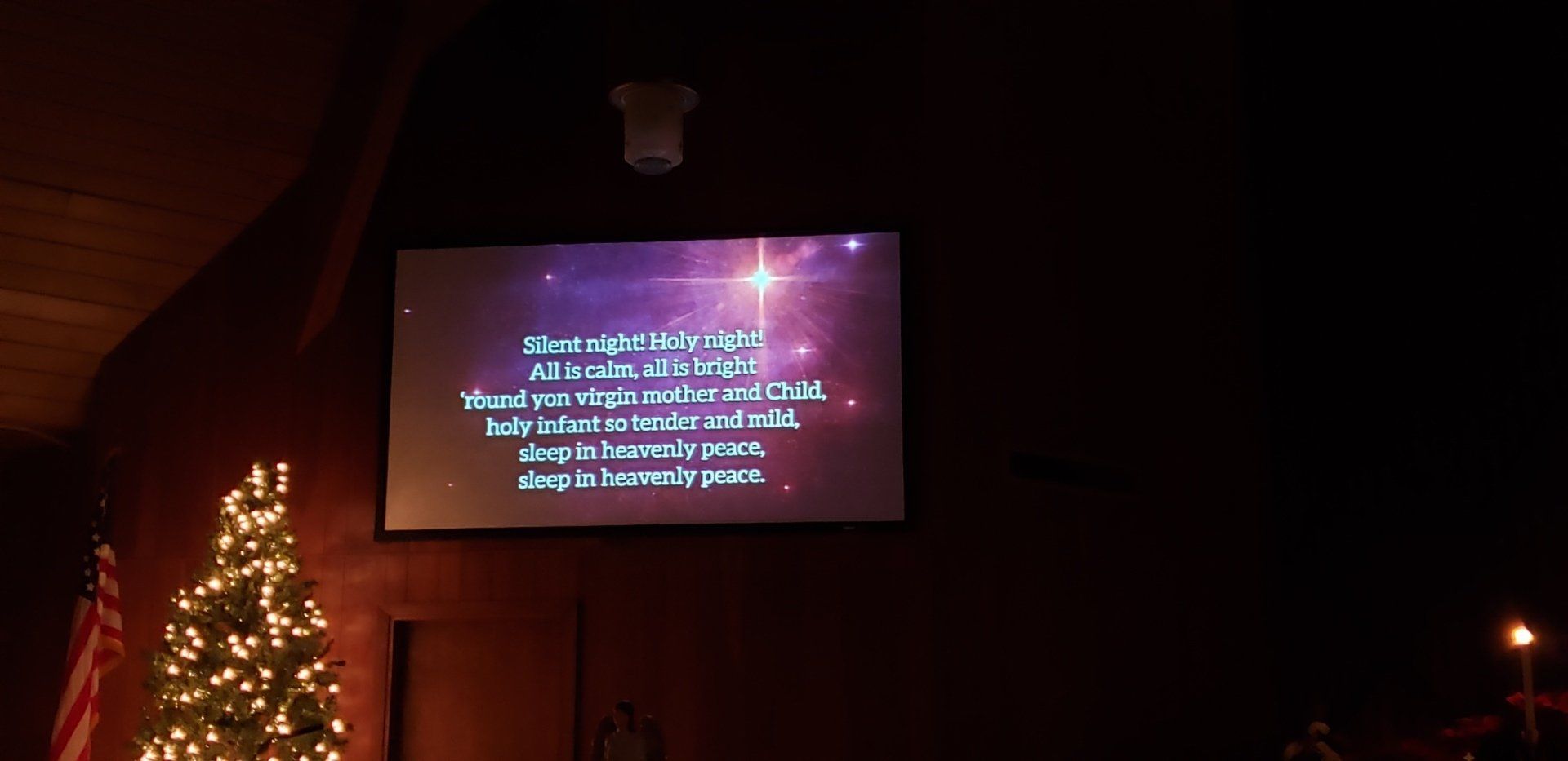 A screen showing white text at night beside a lit Christmas tree and dark building exterior