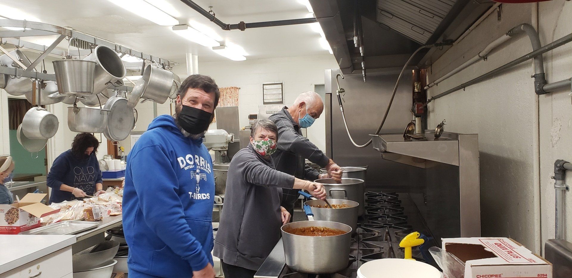 People preparing food in a commercial kitchen, including one in a blue hoodie and mask stirring a pot.