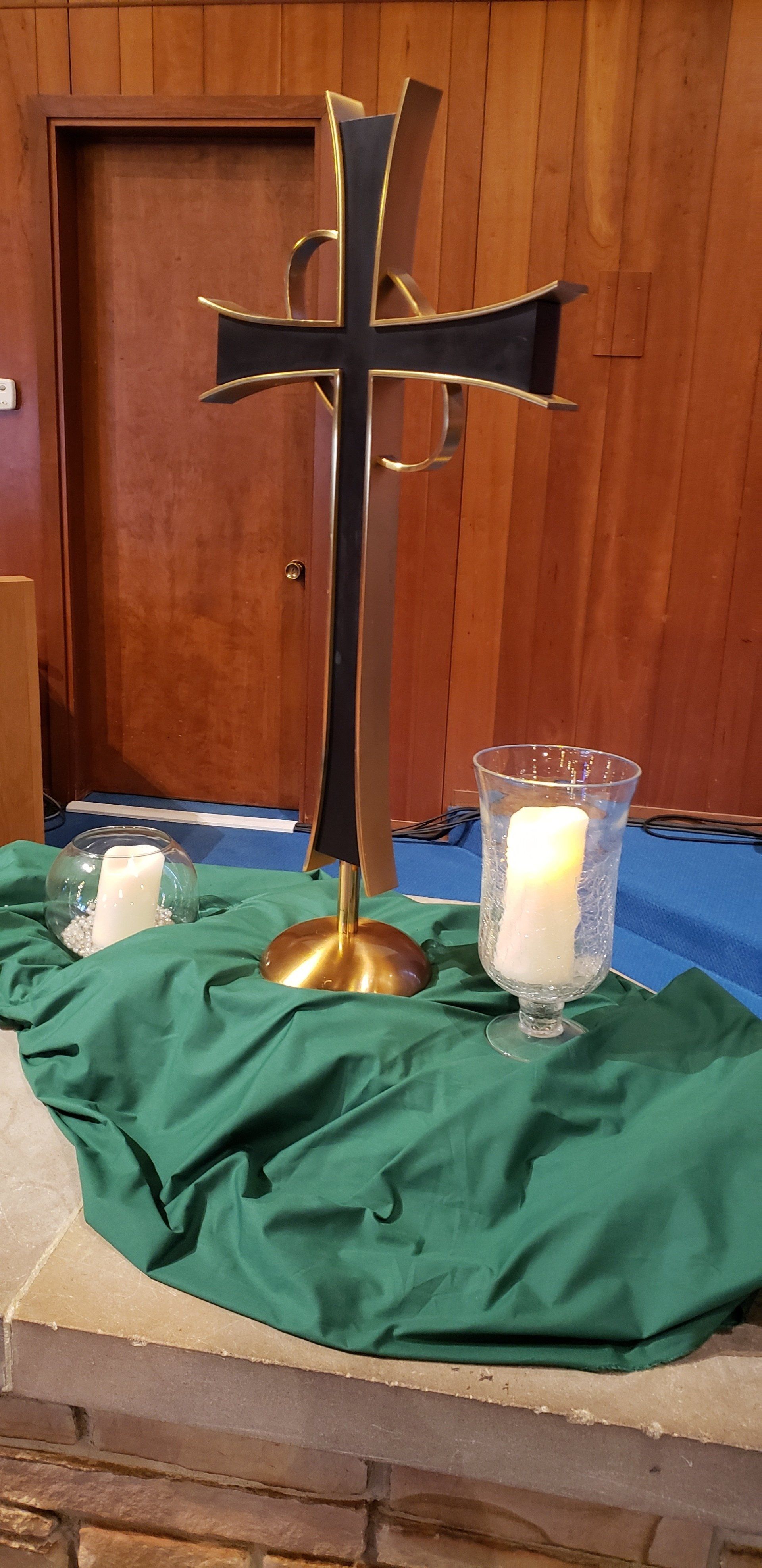 A black cross on a brass stand beside lit candles on a green cloth in a wooden chapel.