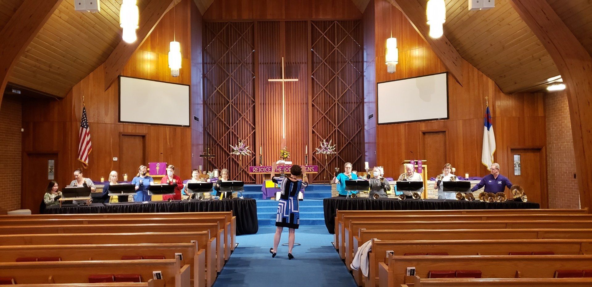 Wide view of a church sanctuary with a choir and congregation seated in pews, wood-paneled walls, and a cross altar
