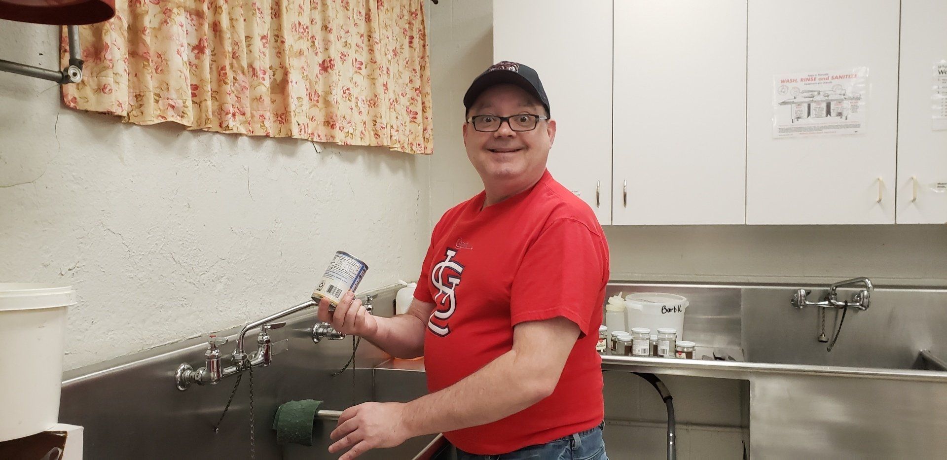 Person in red shirt washing dishes at a sink in a kitchen, smiling at the camera.
