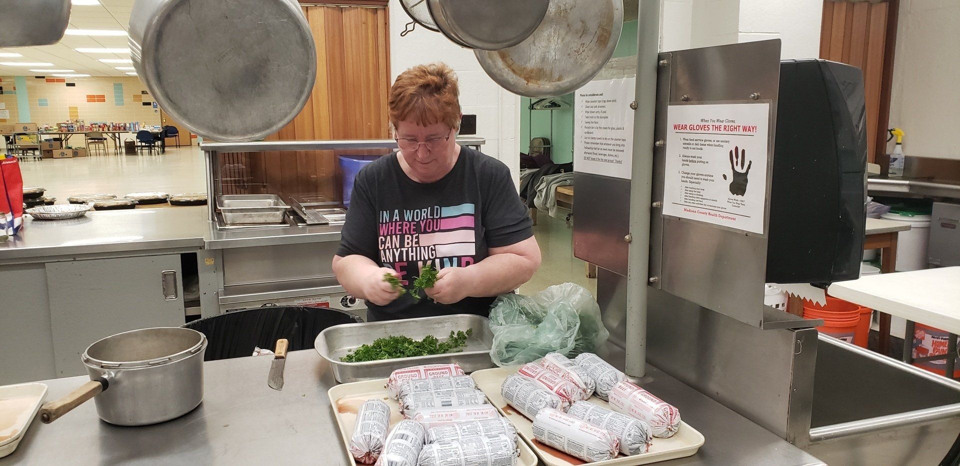 Person preparing food in a commercial kitchen beside trays of wrapped items and large pots