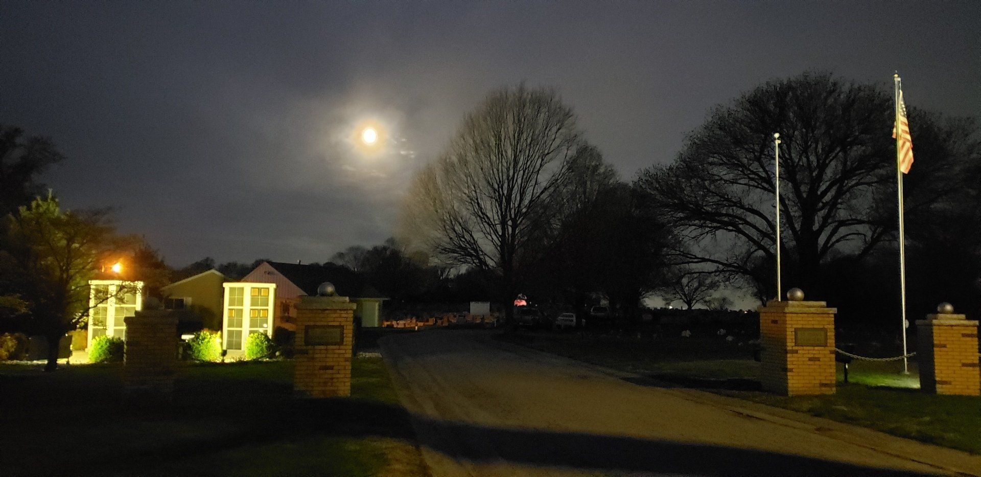 Moonlit driveway entrance with lit brick pillars, trees, and a flag at night