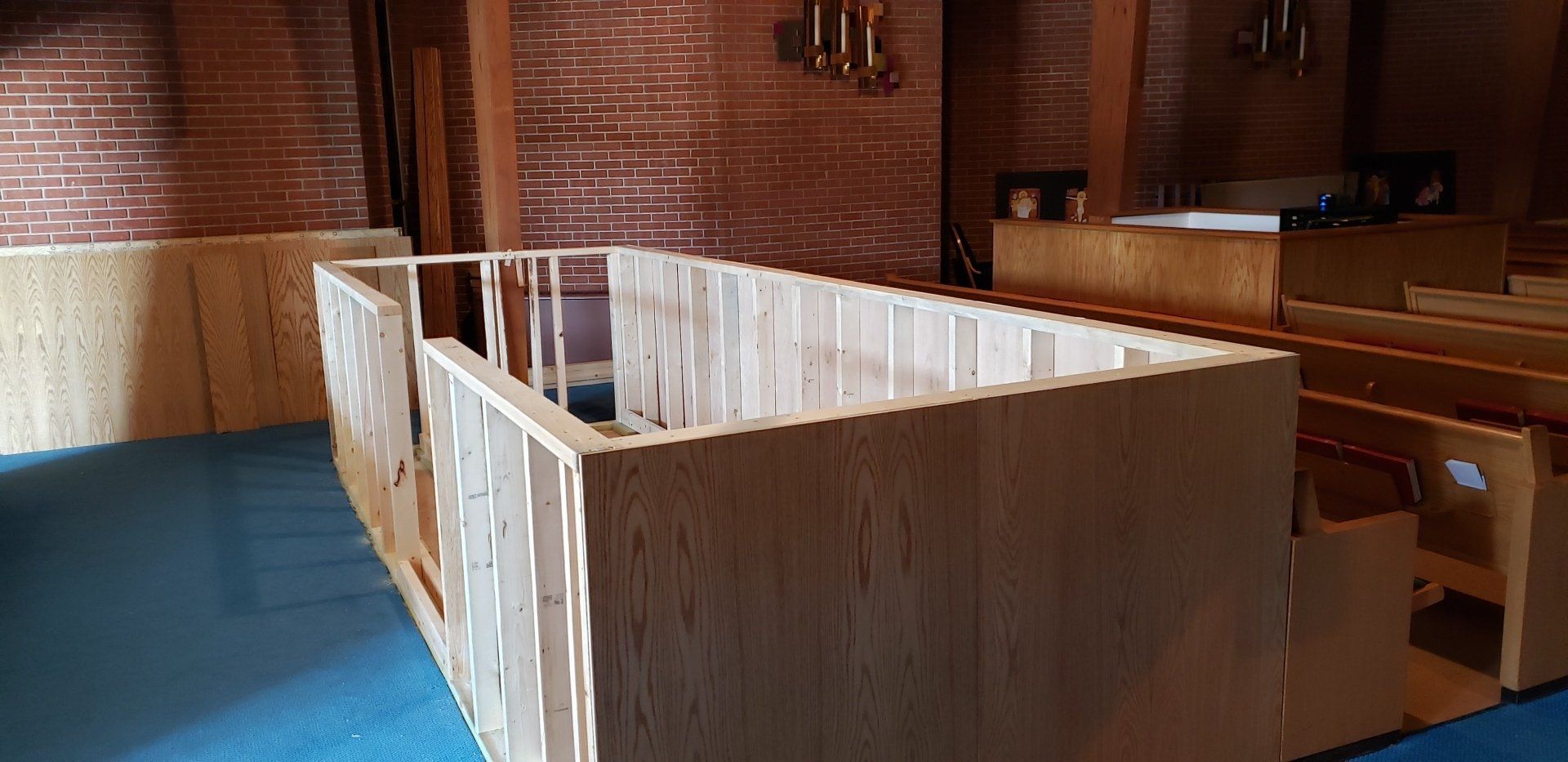 Wooden fenced enclosure inside a brick-walled church or hall with blue carpet and pews nearby