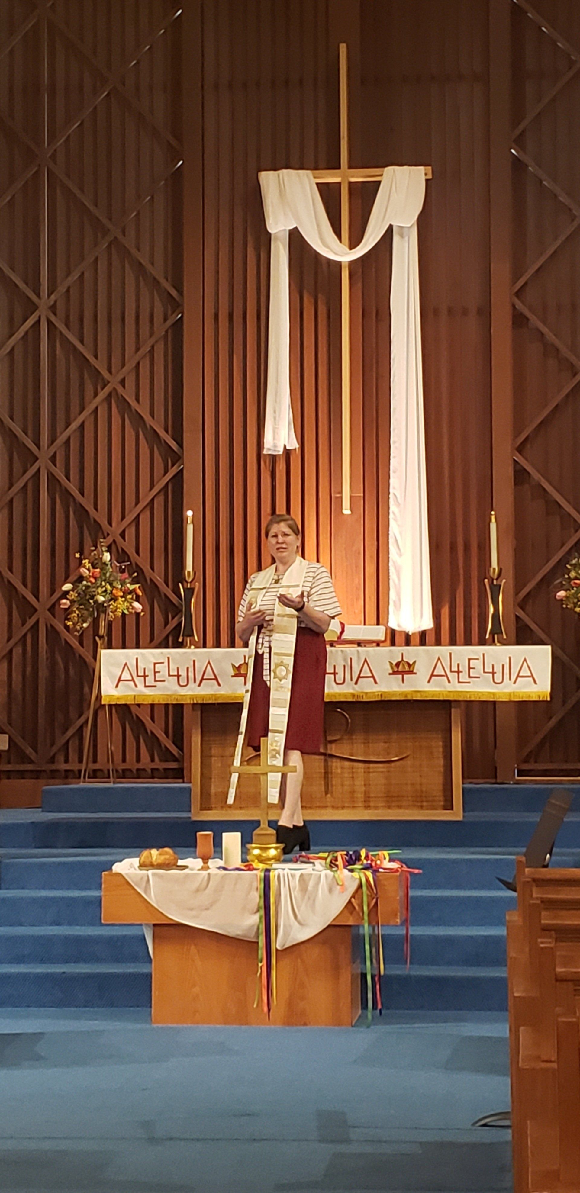 Person speaking at a church podium before a cross draped in white fabric, with blue steps and altar decor
