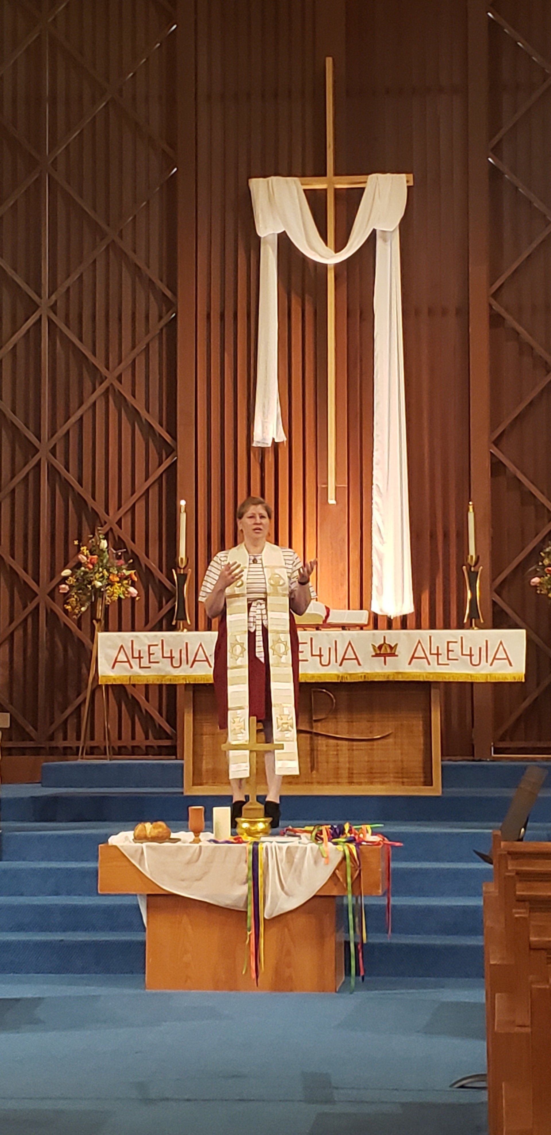 Person speaking at a church pulpit beneath a white drape and cross on a blue-carpeted stage