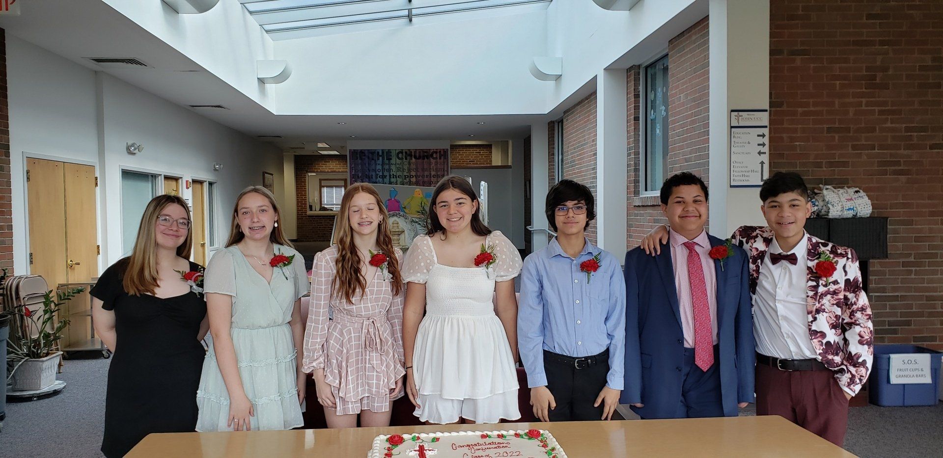 Seven people stand together indoors, smiling in formal attire at a celebration or event.
