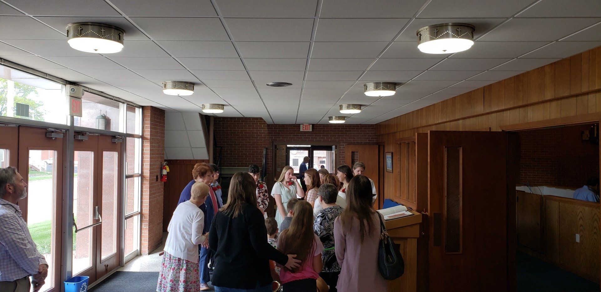 People gathered in a hallway under round ceiling lights, with wood-paneled walls and glass doors.
