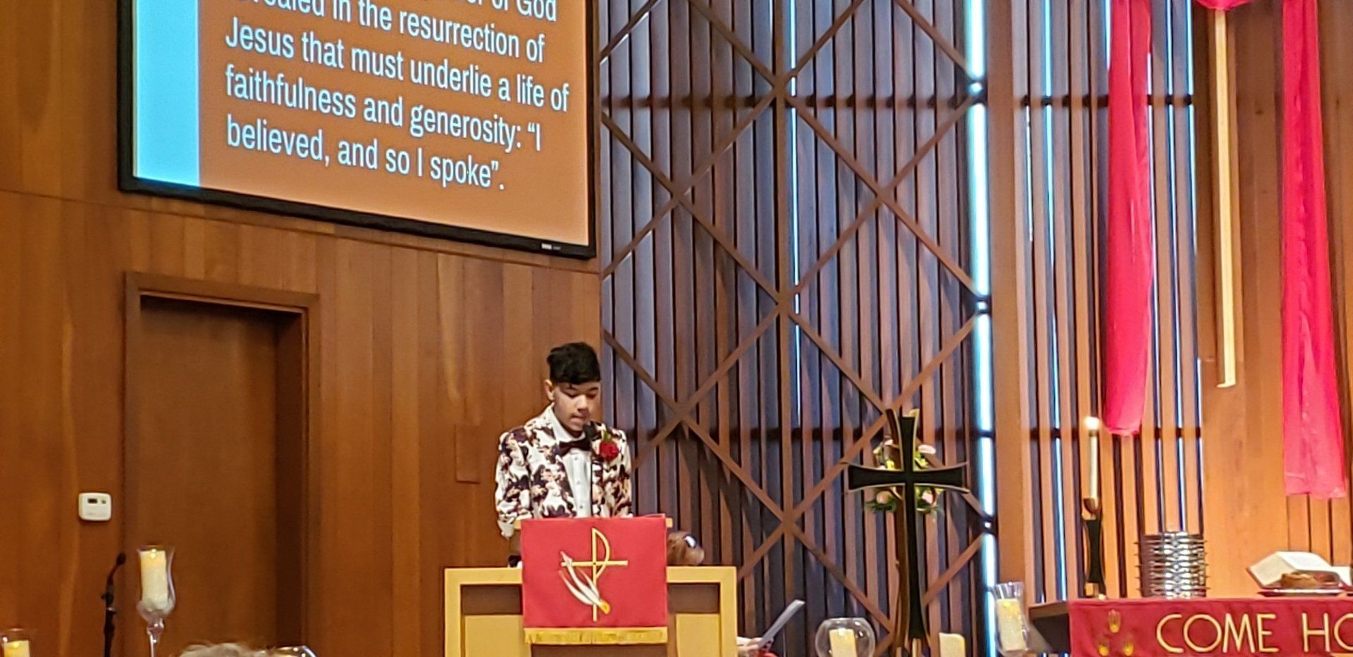 Speaker at church podium under a cross, with projected text and red drapery in the background