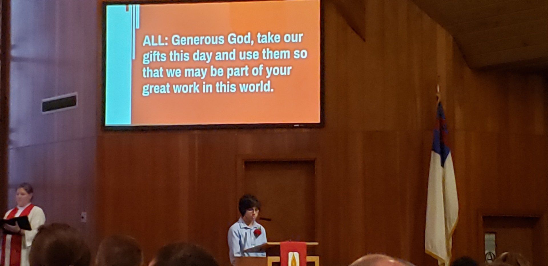 Person speaking on a stage beneath a projected Bible verse, with podiums and flags in a church auditorium