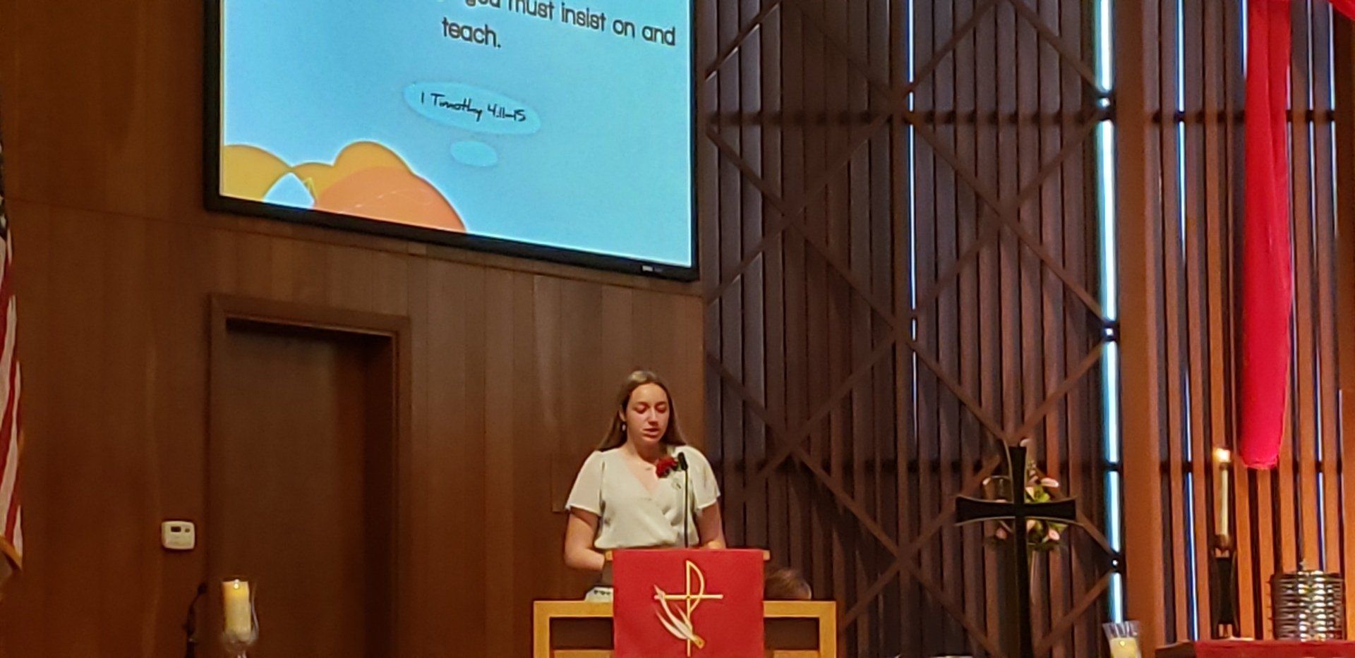 Speaker at a podium in a church, with a projected slide and red flag backdrop.