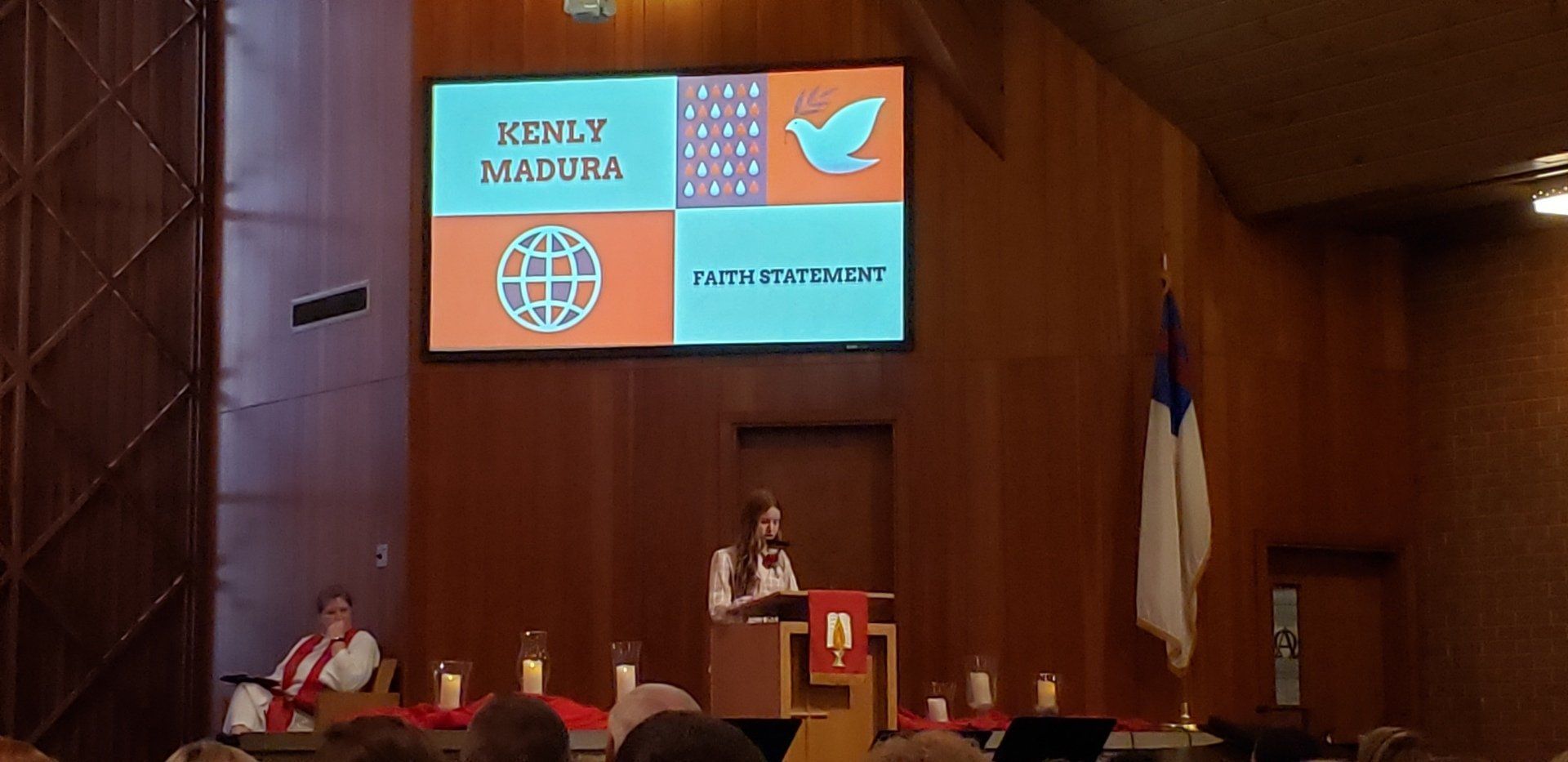 Speaker at podium in wood-paneled hall, with projected presentation screen and seated panelists behind.