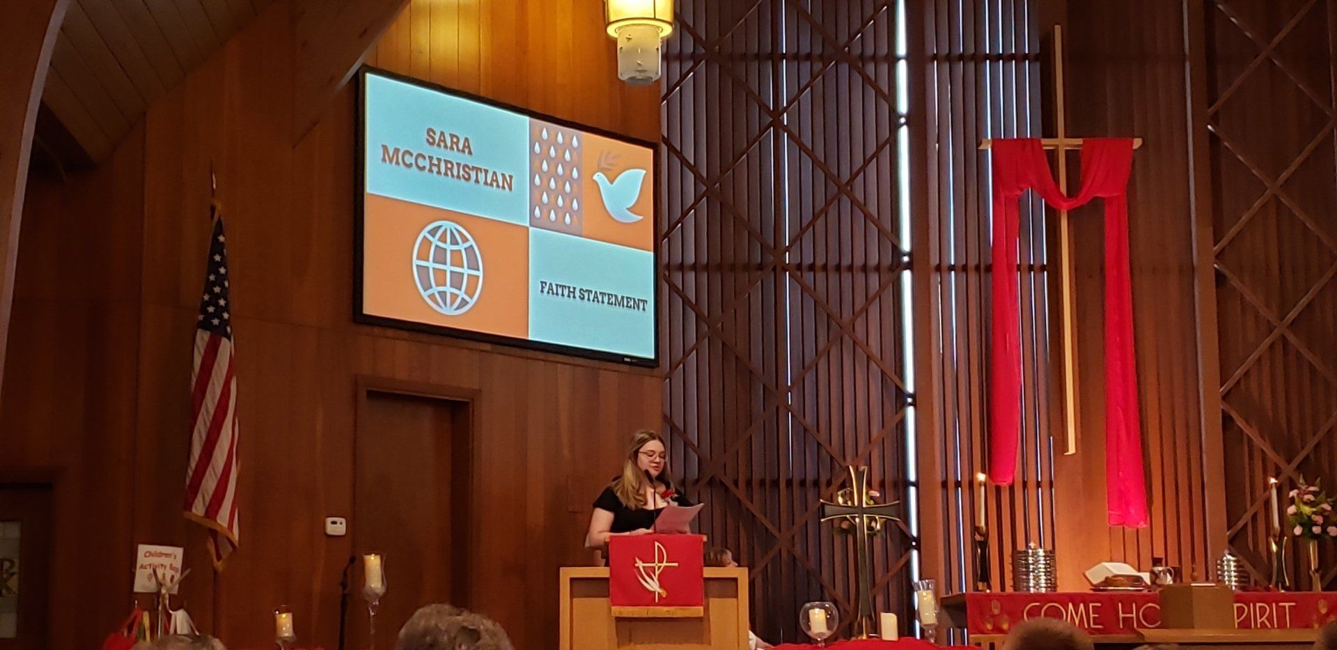 Speaker at a church pulpit with red banners, American flag, and projected slides in a wood-paneled sanctuary