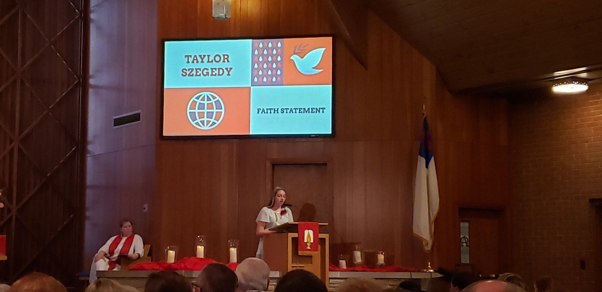Speaker at podium in wood-paneled hall, with projected event slides and seated panelists behind them.