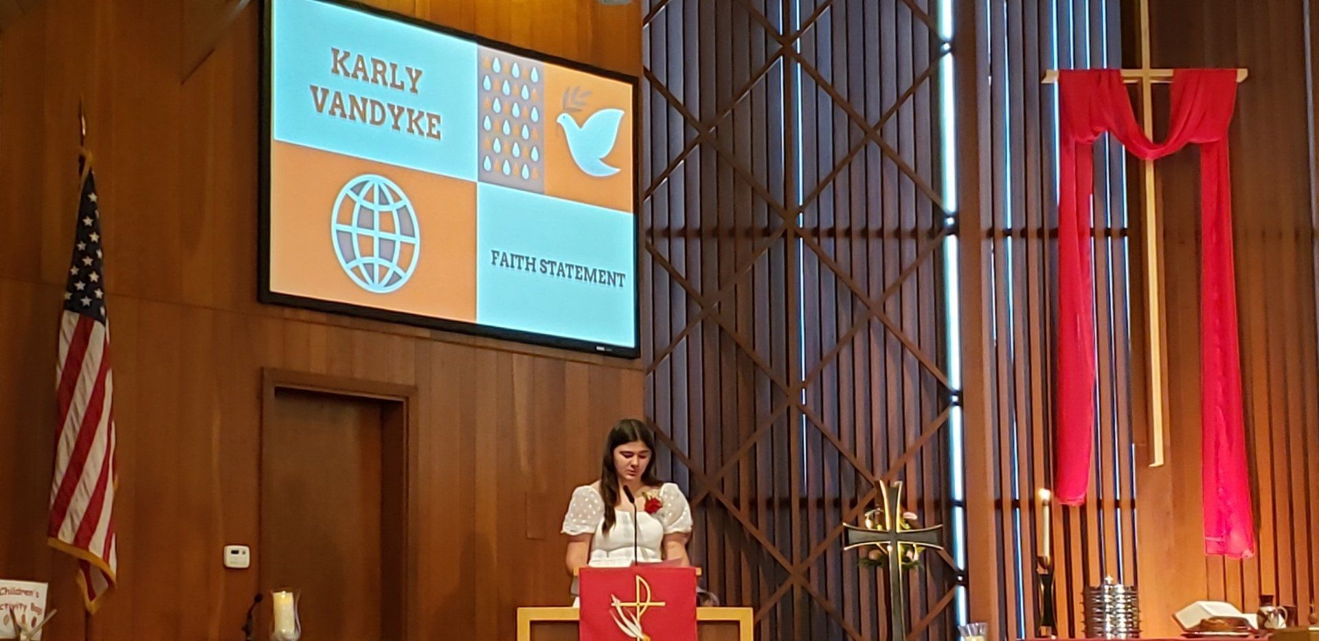 Speaker at podium in wood-paneled auditorium with large presentation screen and red banners behind her