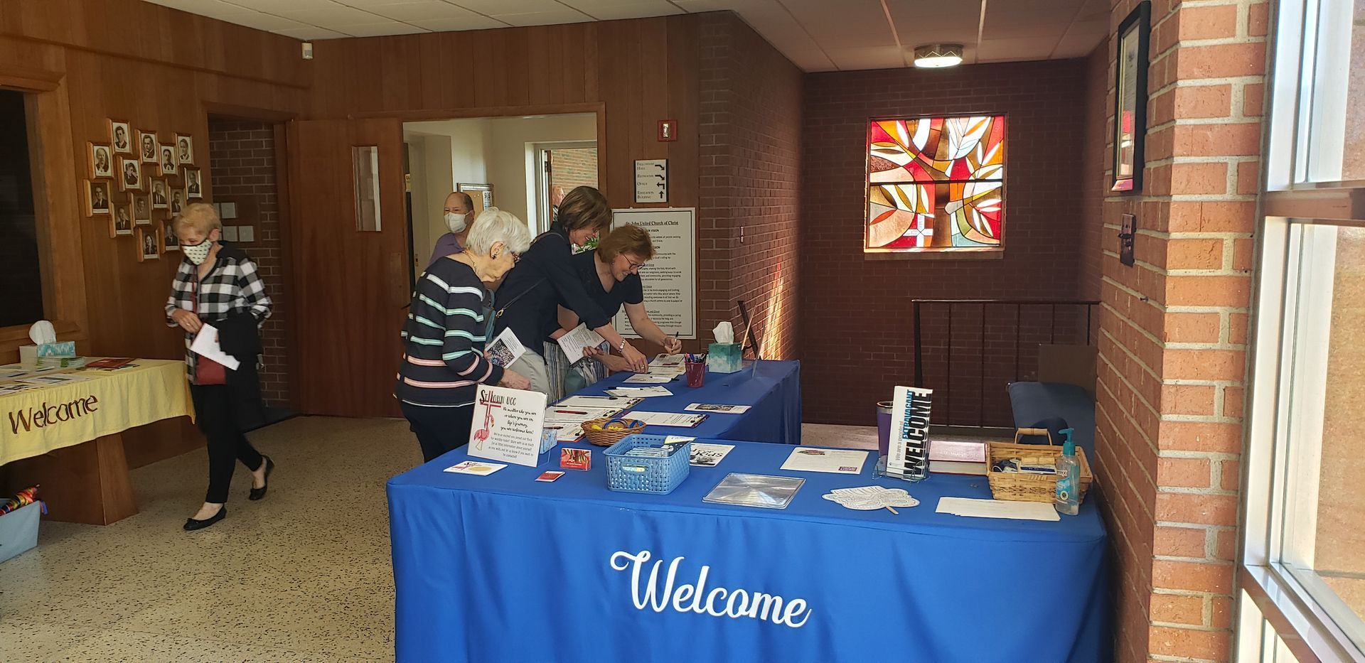 Welcome table in a brick hallway with volunteers standing nearby and a colorful stained-glass window.