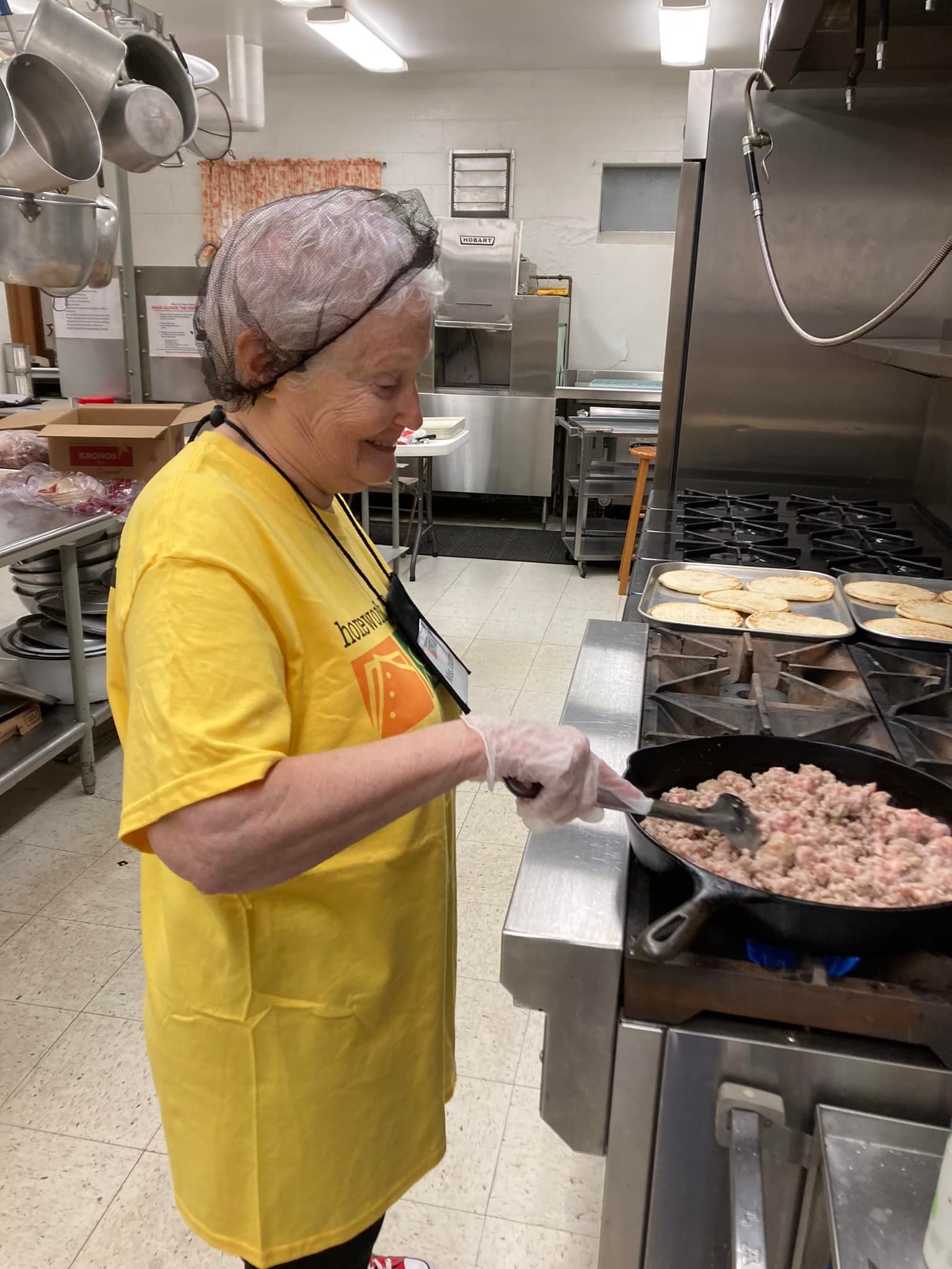 Worker in a yellow apron and gloves cooking meat on a griddle in a commercial kitchen