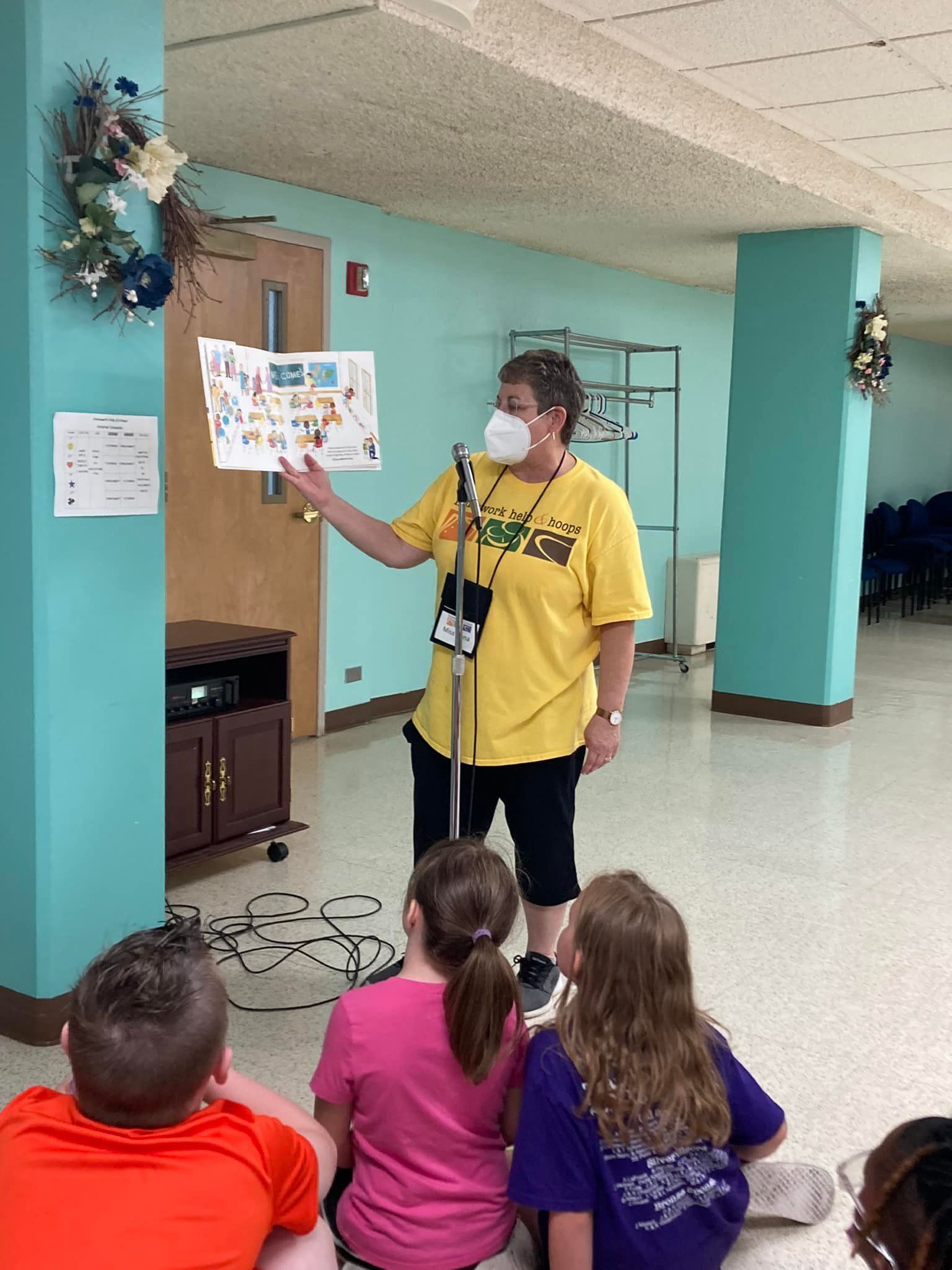 Speaker in yellow shirt points to poster for seated children in a hallway.