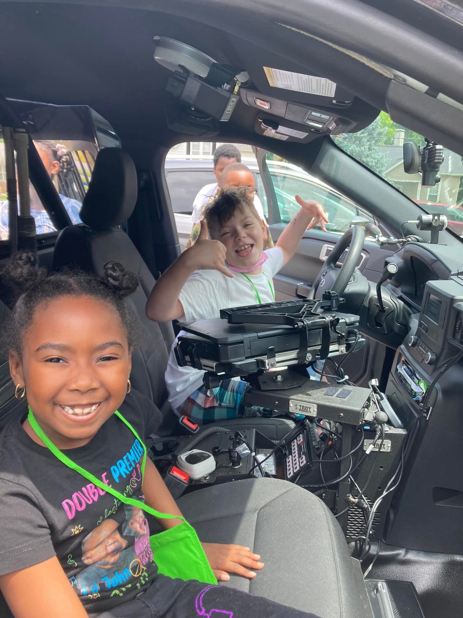 Two children smiling inside a car, one in the front seat with a green strap, one in the back waving.