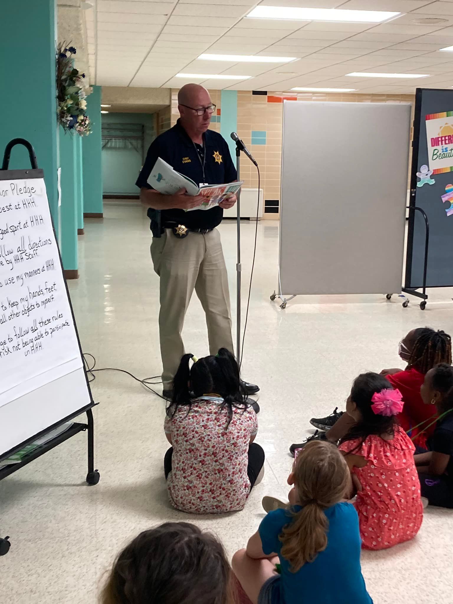 Instructor reading to seated children in a bright classroom hallway, with flip charts and a whiteboard nearby