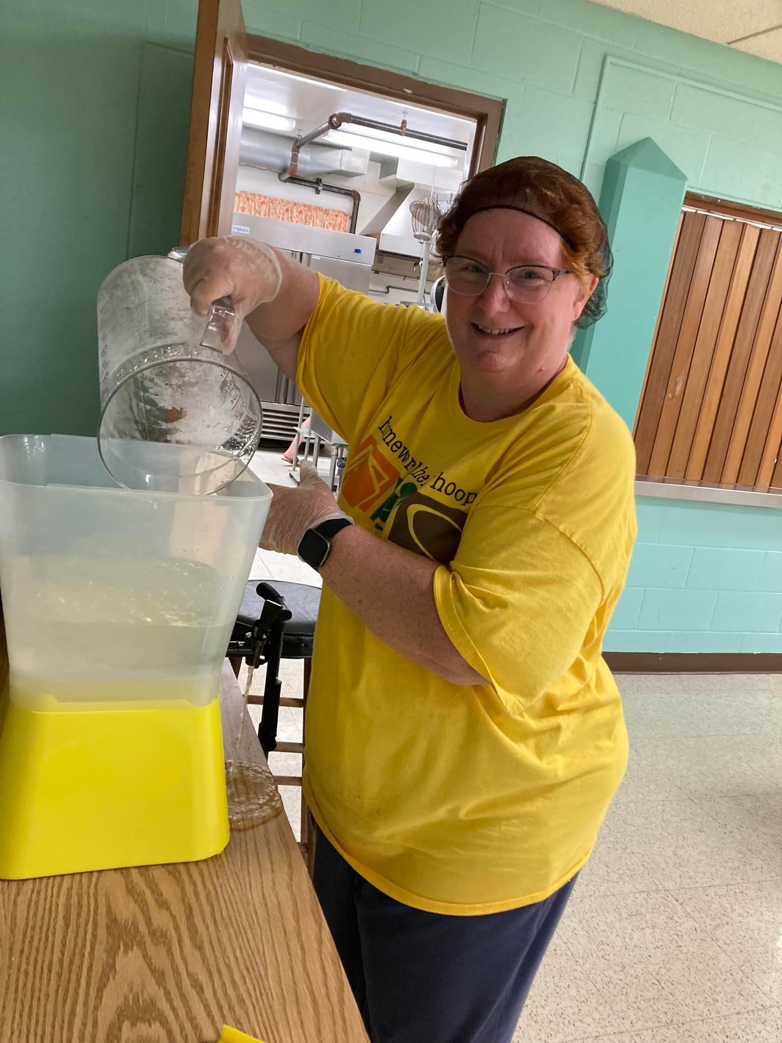 Woman in yellow shirt pouring batter into a container in a kitchen, smiling.