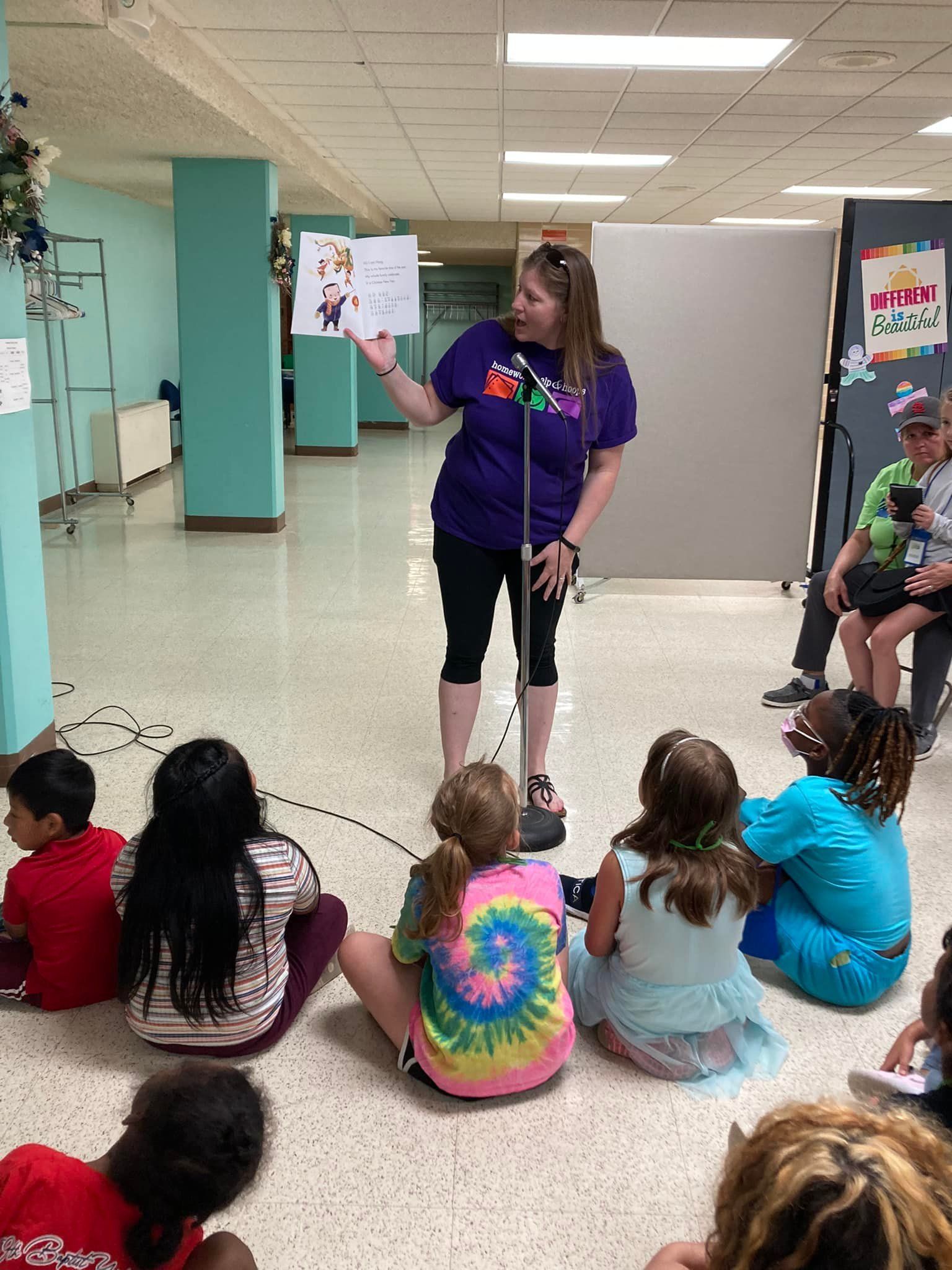 Woman in purple speaks to seated children in a hallway, pointing to a poster on a display board.