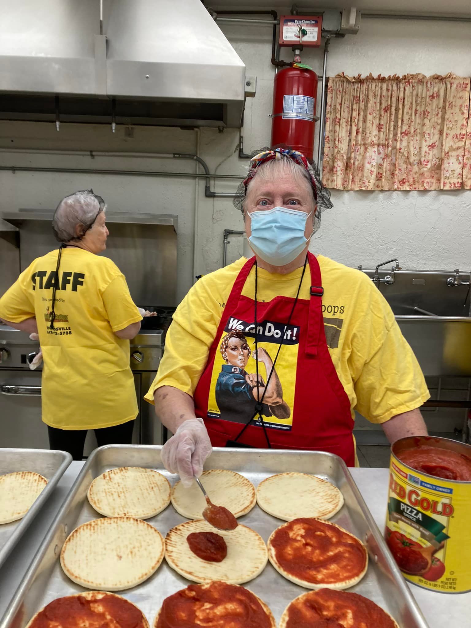 Volunteer in red apron and mask arranging pizza dough in a kitchen beside trays and a sauce can