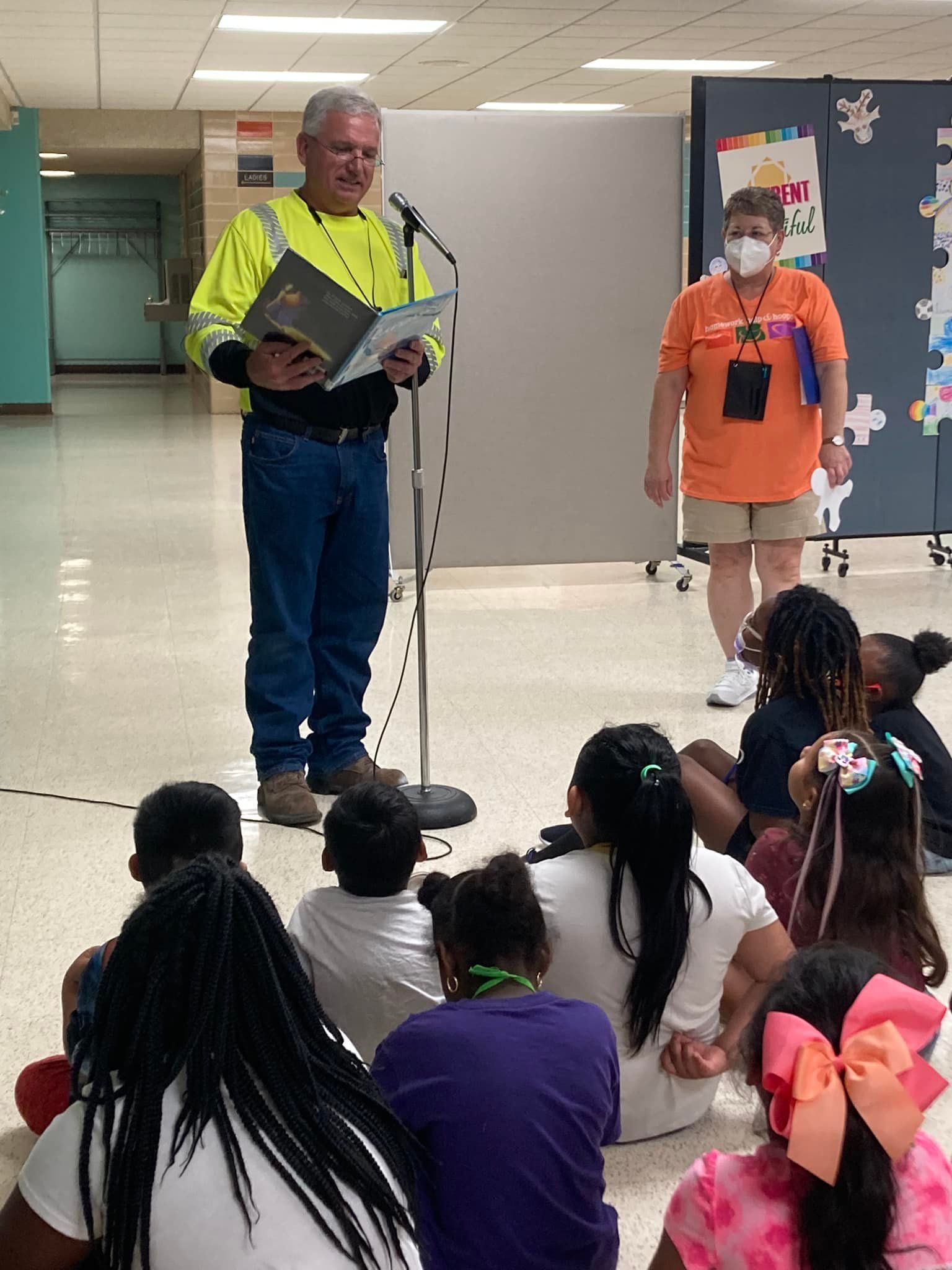 Man in neon safety shirt speaks at a microphone to seated children in a school hallway, with a staff member nearby