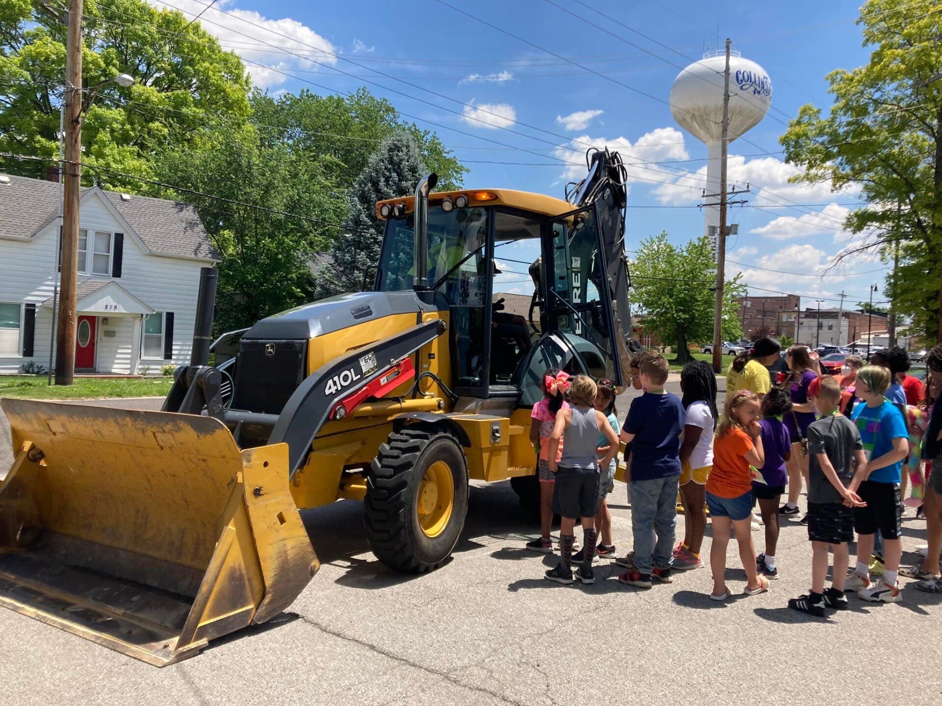 Construction vehicle on a street as a crowd of children gathers beside it under a blue sky.