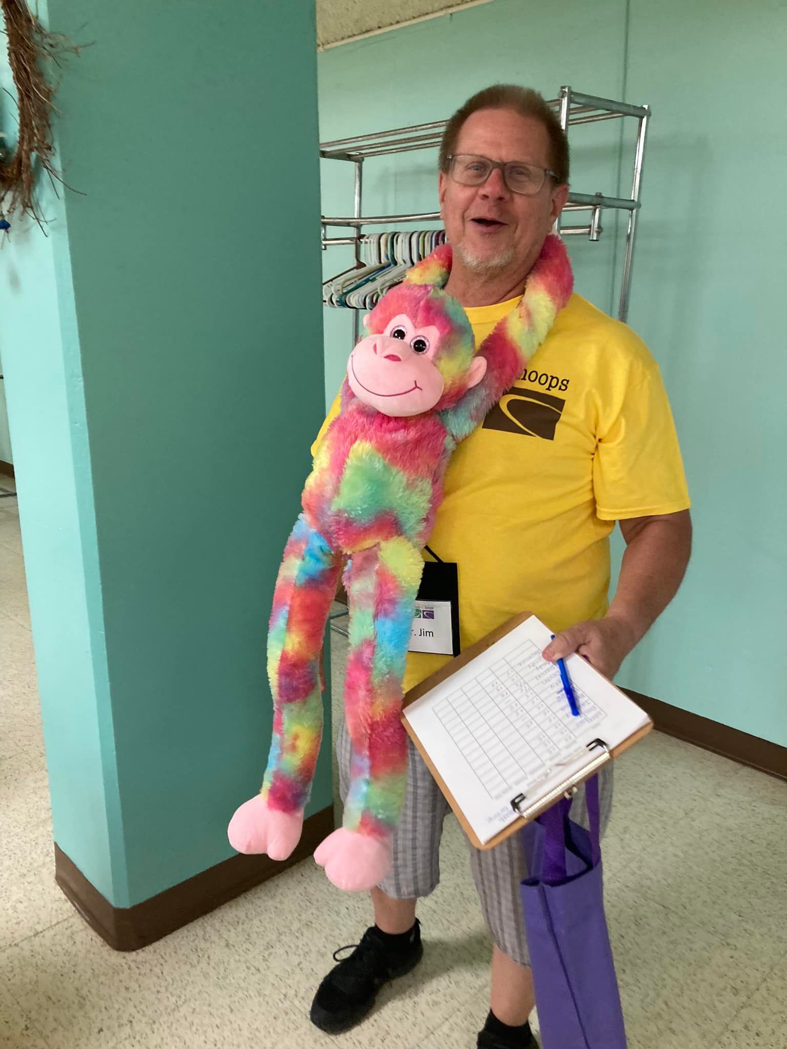Man in yellow shirt holding a rainbow plush toy and clipboard in a hallway