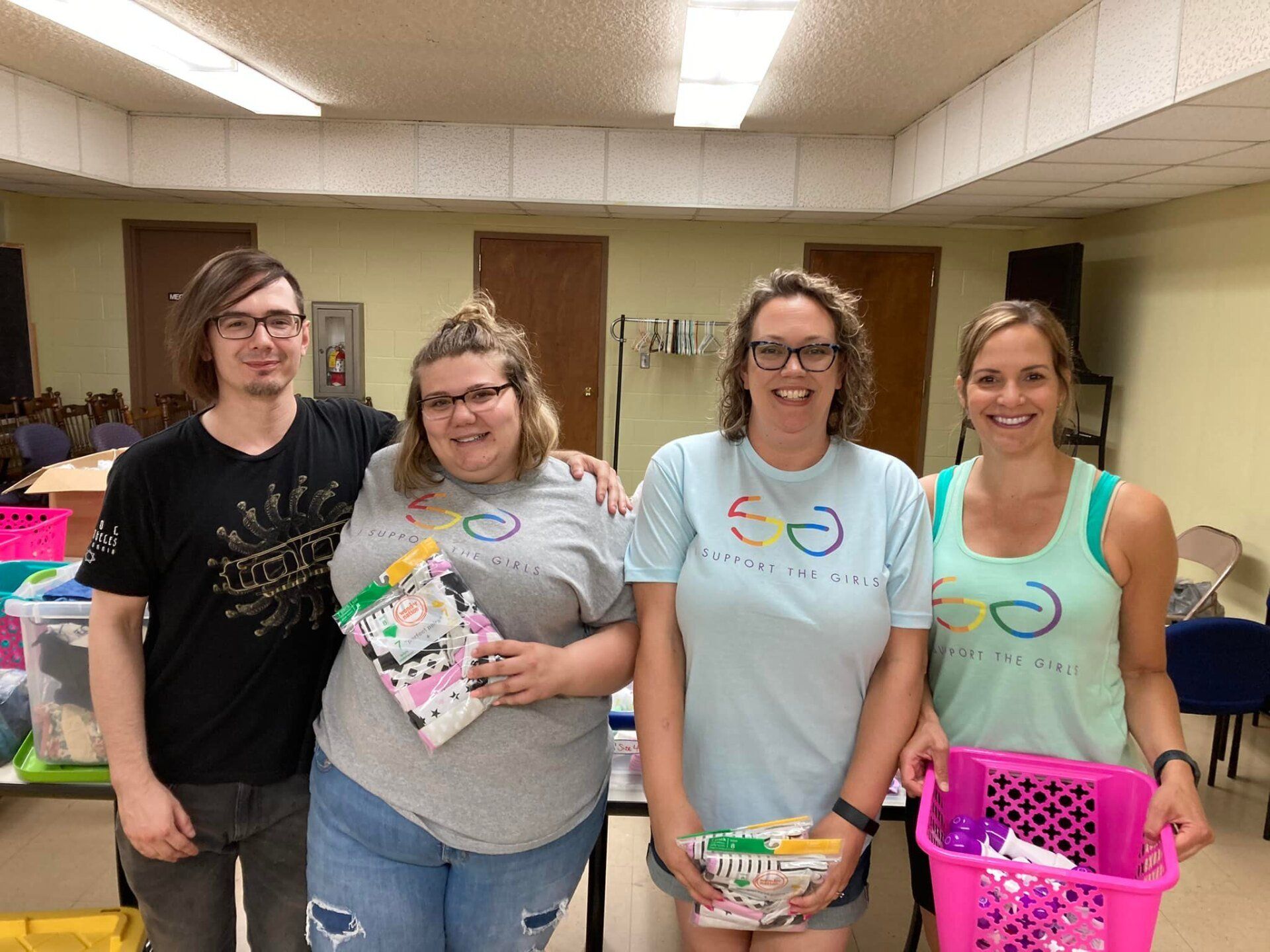 Four people pose indoors with baskets and supplies, smiling in a room with tables and fluorescent lights.