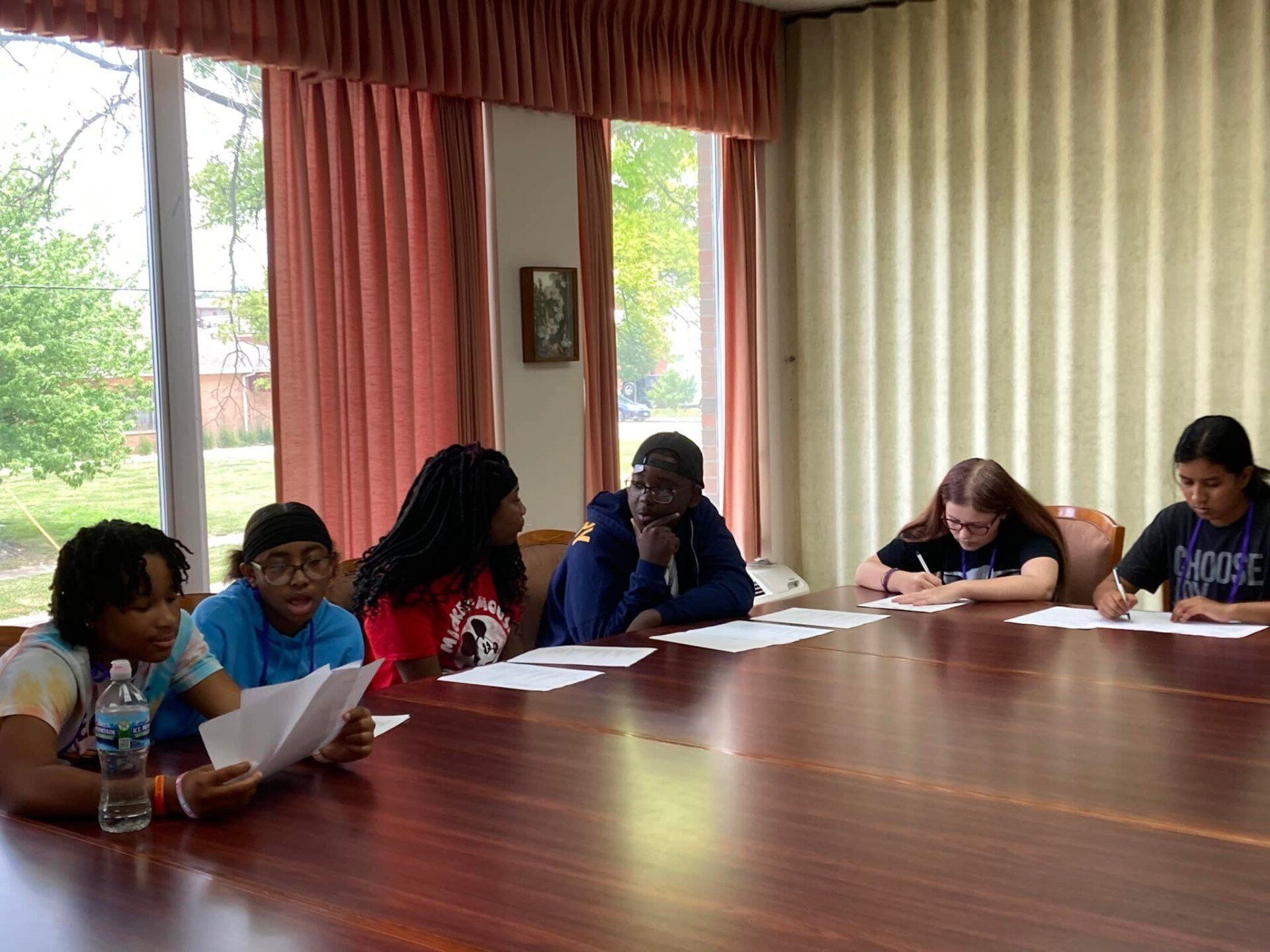 Students seated around a large conference table in a bright meeting room, writing on papers.